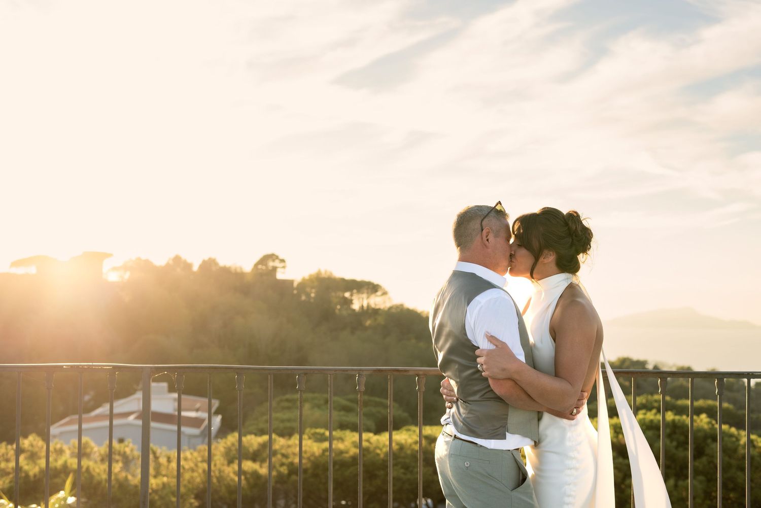 Bride and groom embrace tightly and share a passionate kiss. A soft glow of evening sun peeps out from the clouds onto surrounding trees.
