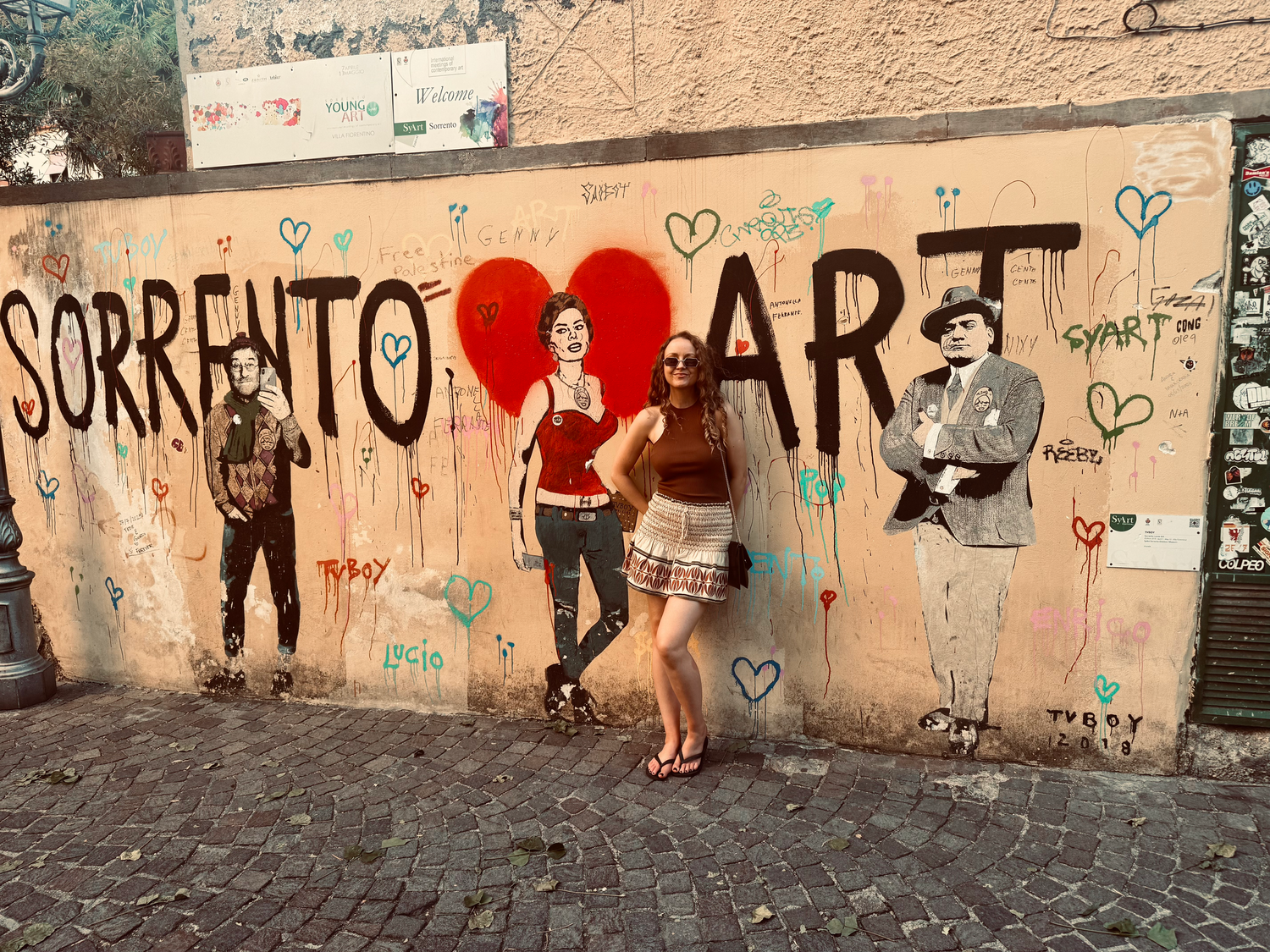 Woman leaning against a wall decorated in street art. Lettering on the wall says Sorrento Art and there is a mural of a woman in a heart.
