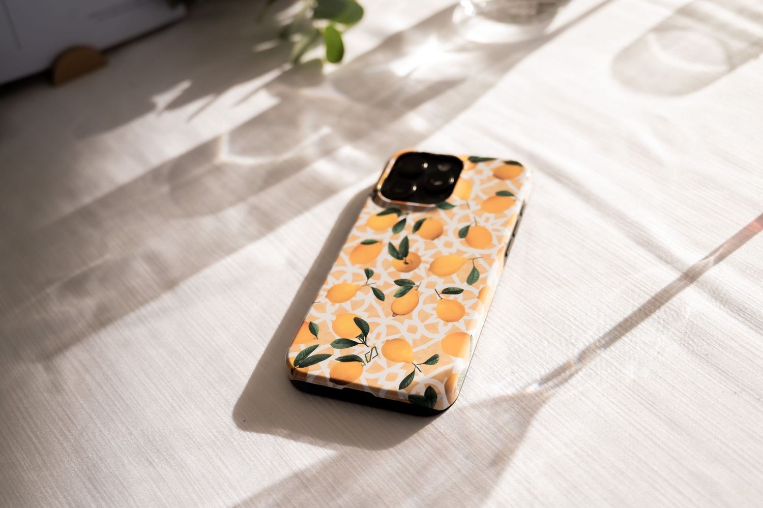 I phone on a table in the sunlight in a yellow phone case, the case design has big and small lemons with leaves dotted on it.