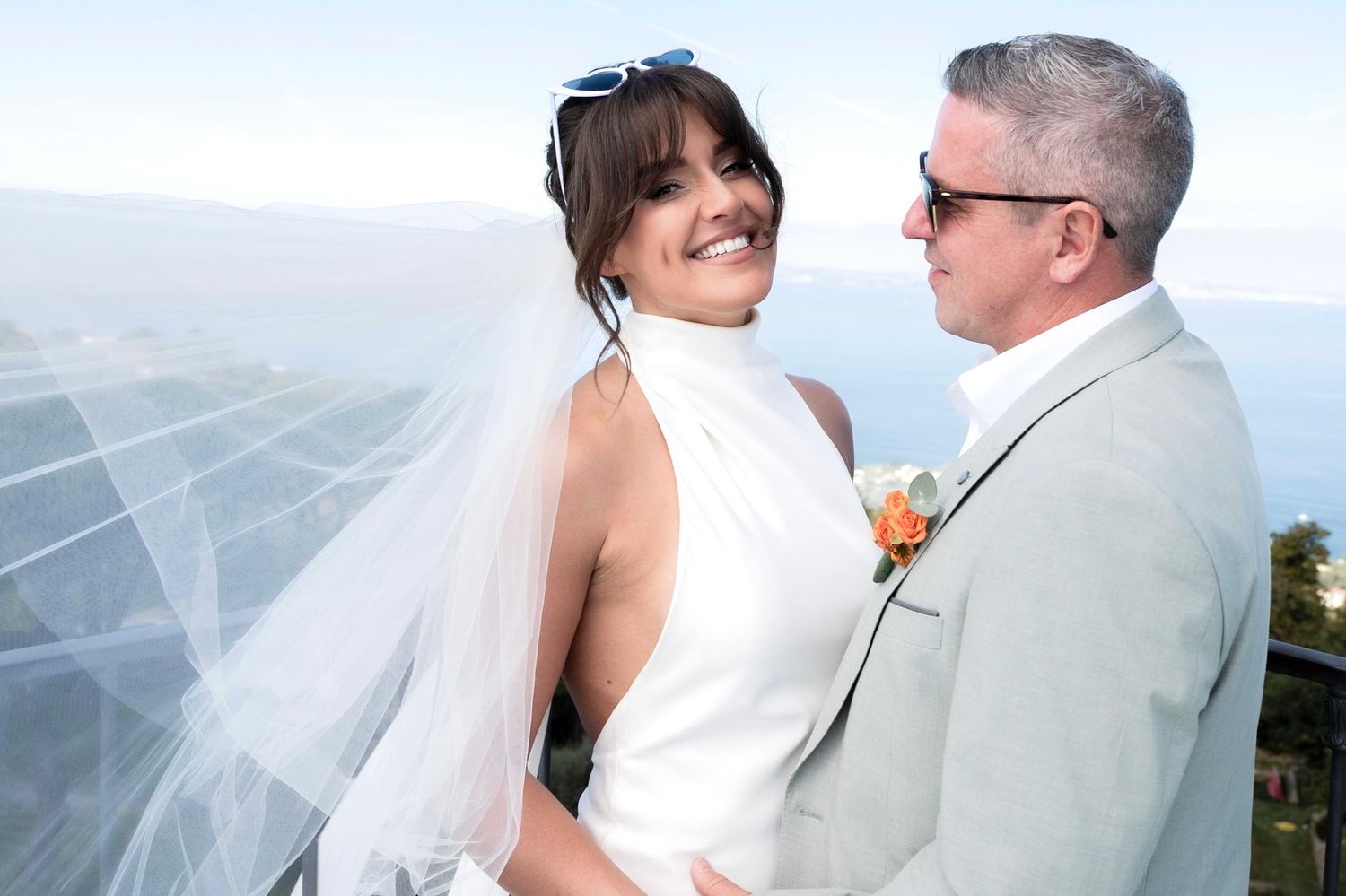 Husband admiring his beaming bride as they stand by the ocean and her veil flows behind her in the wind.