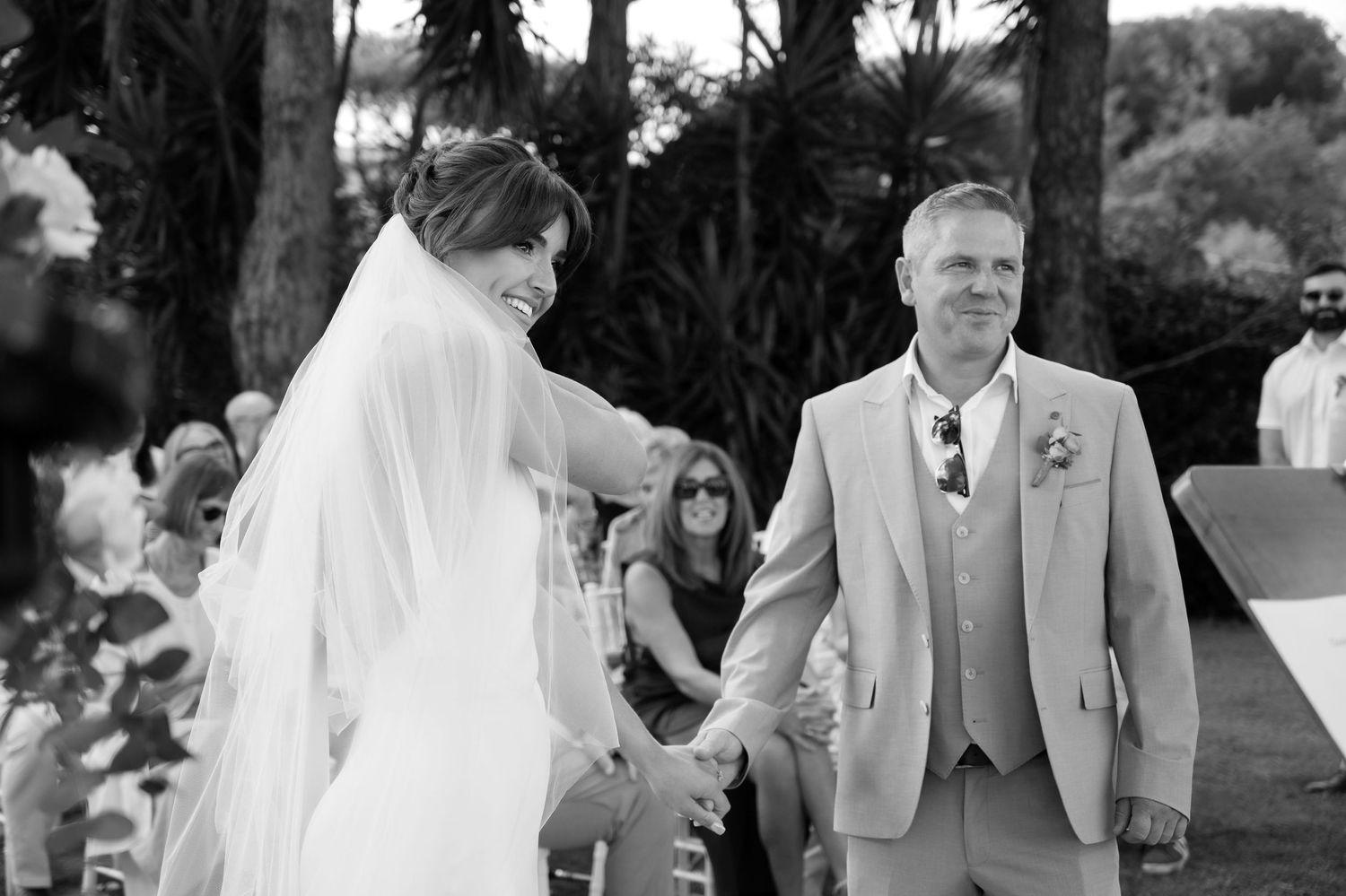 Newly weds holding hands at the alter in a tree lined garden whilst the smiling guests look on. The bride is adjusting her veil.