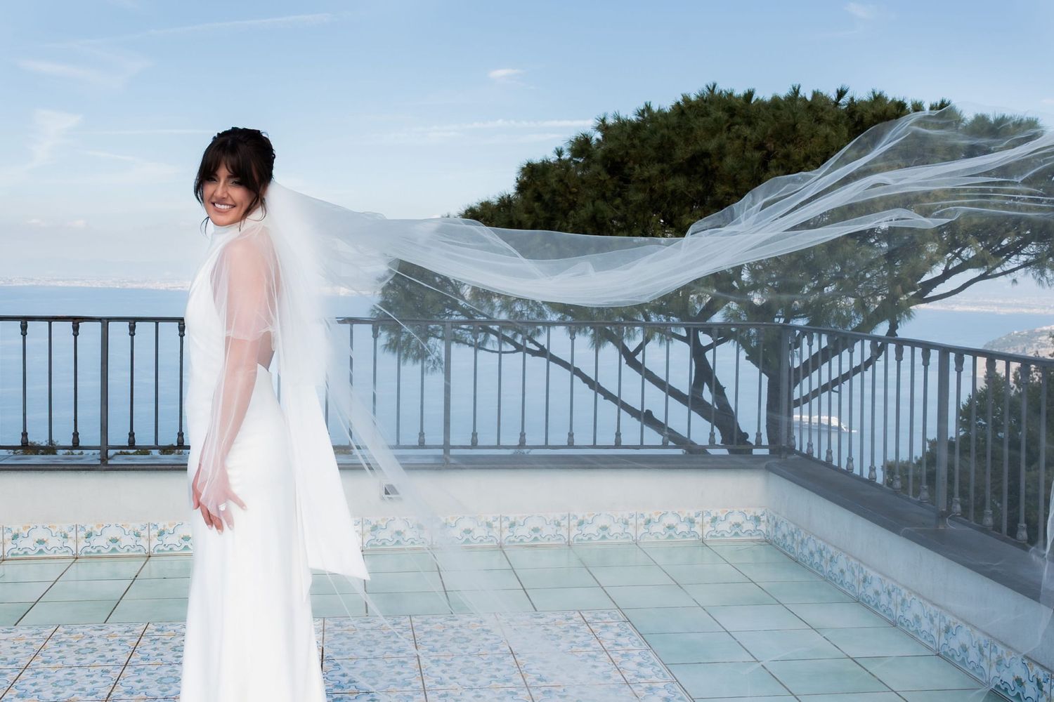 Smiling bride standing on a rooftop terrace on the coast with her hands by her sides and her wedding veil flowing in the wind behind her.