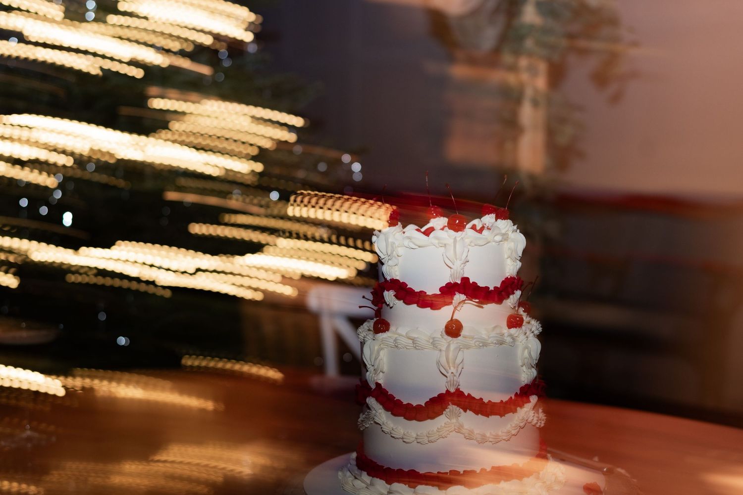 A two tiered ruffle cake decorated with glacier cherries sits elegantly on a wooden table as party lights swirl around it.
