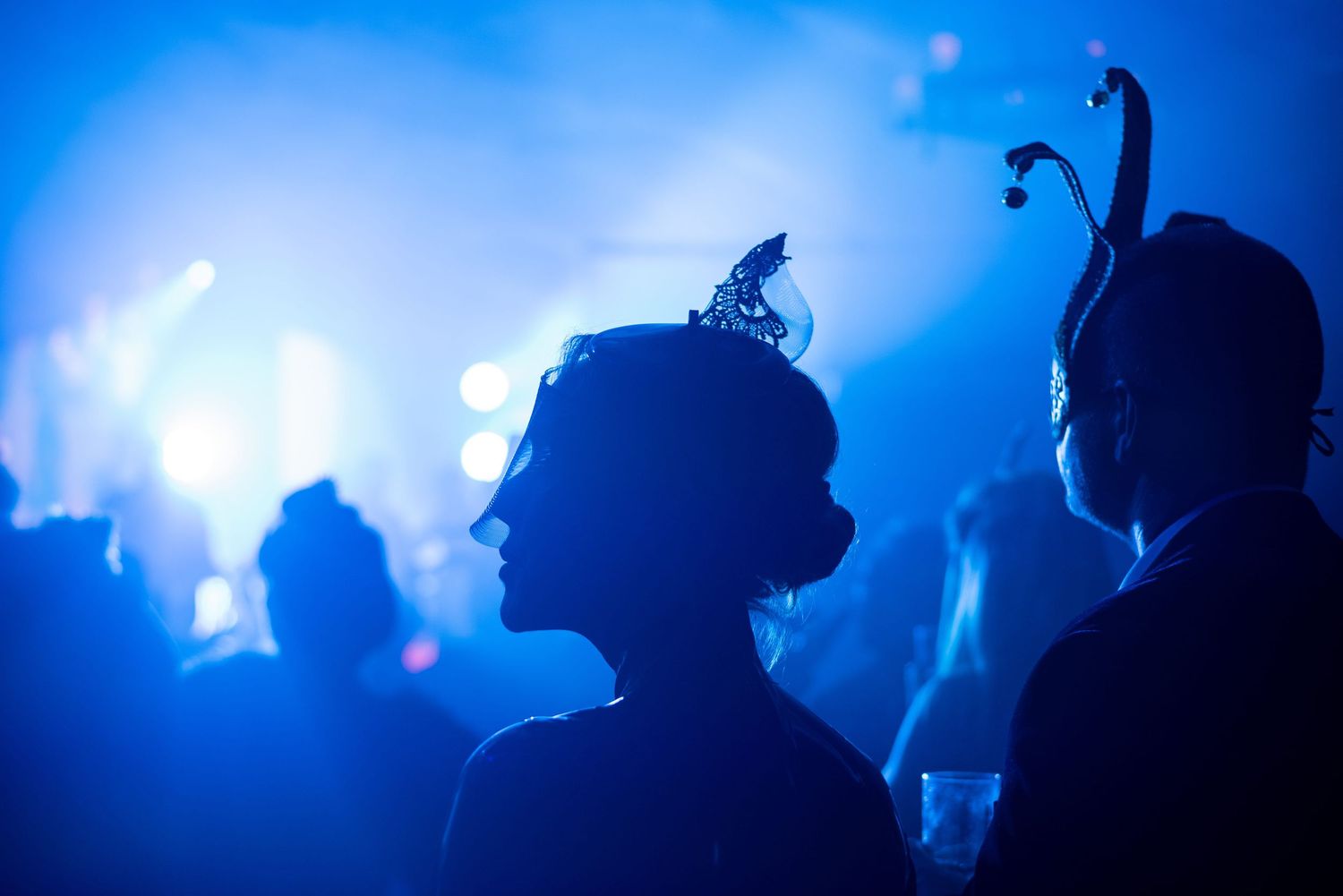 Silhouette of smartly dressed partygoers at a masquerade ball wearing elaborate lace and bell decorated headpieces.