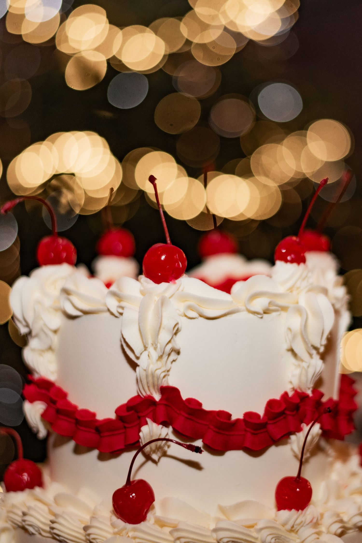 The top tier of a white and red ruffled wedding cake, topped with shiny red glacé cherries with long stalks. Gold fairy lights create bokeh.