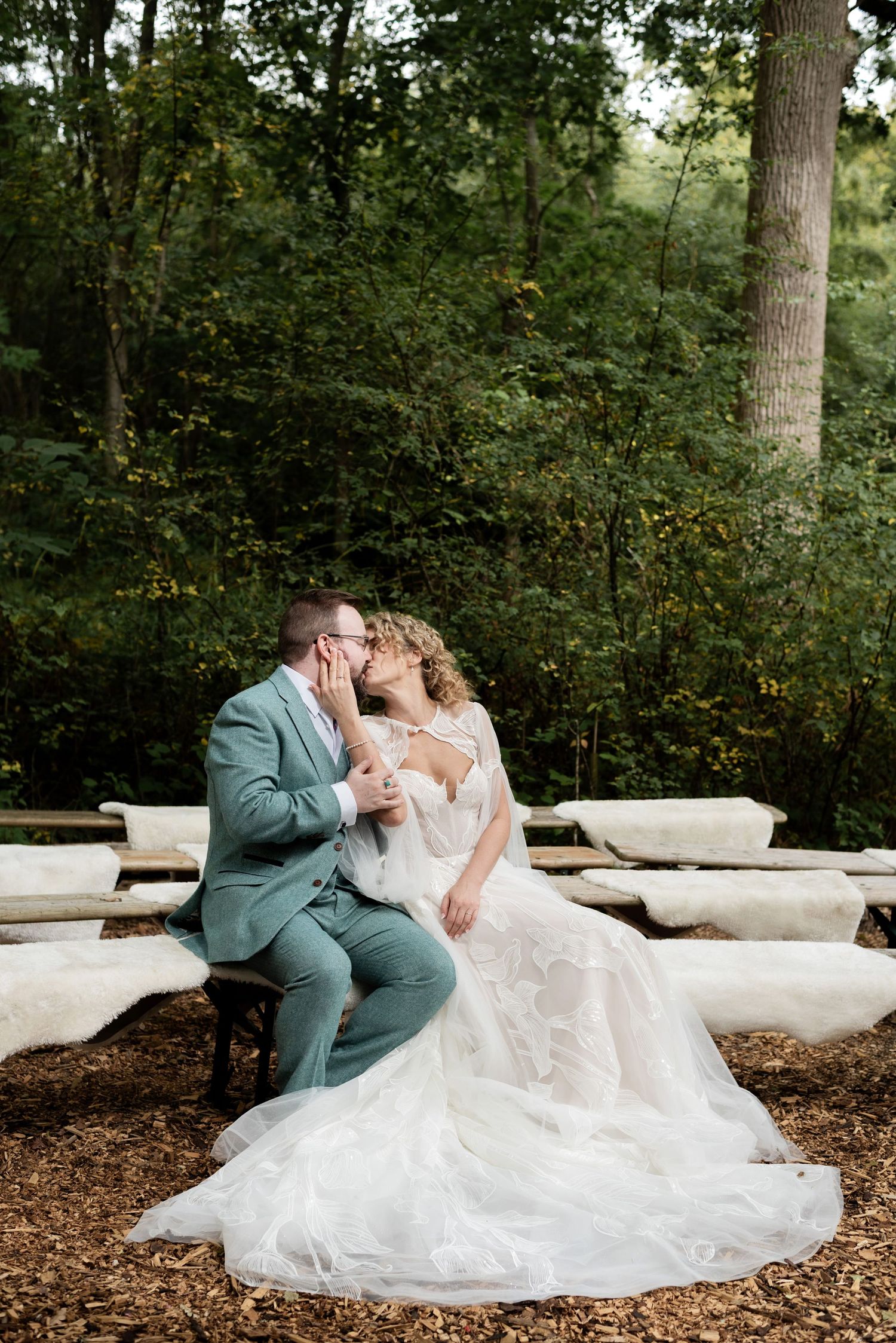 A couple sit closely on wood covered blankets in the forest sharing a kiss. The woman holds one arm up and cups the mans face in her hand.