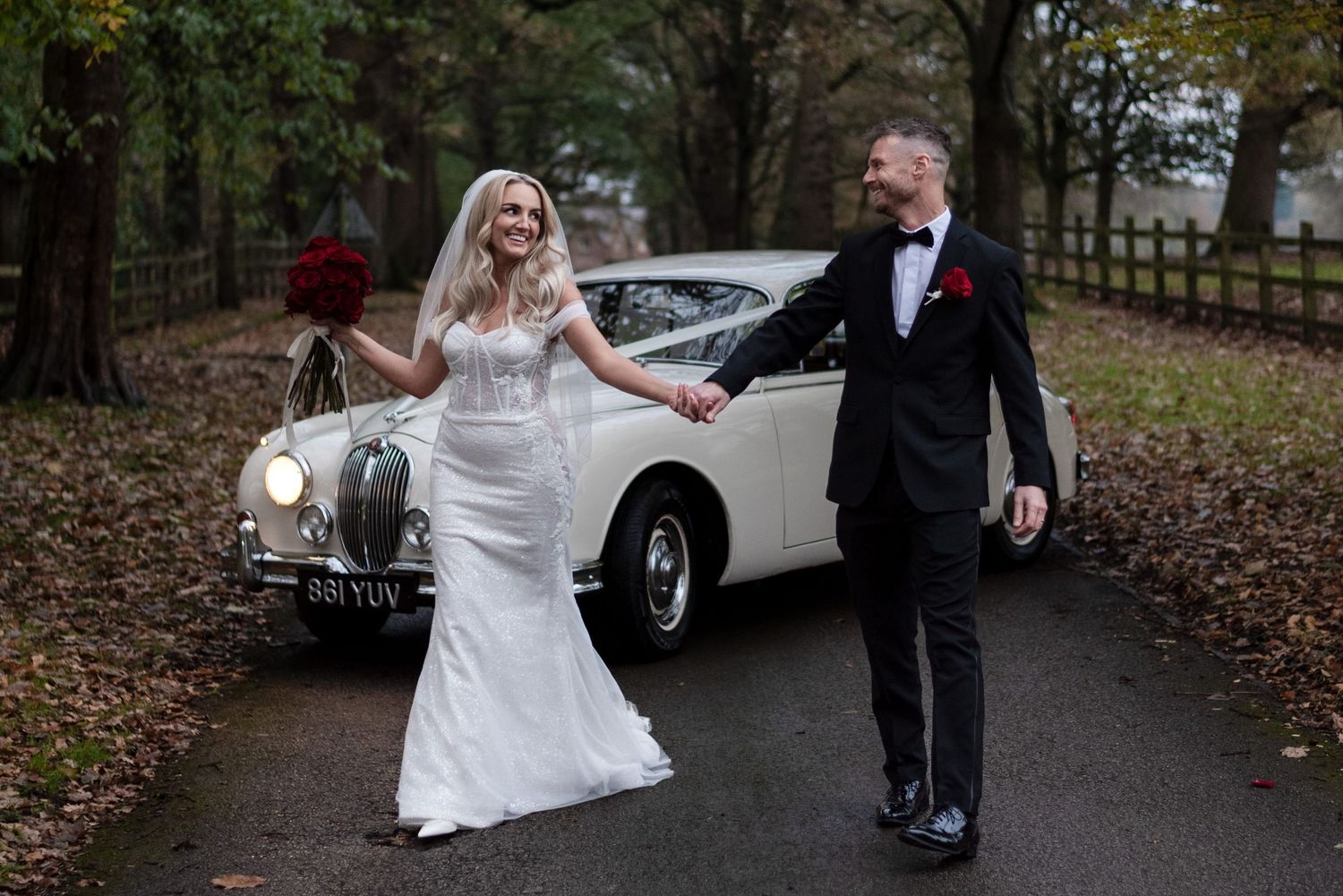 A newly married couple laugh together joyously as they walk hand in hand down a tree lined driveway, their classic car parked behind them.