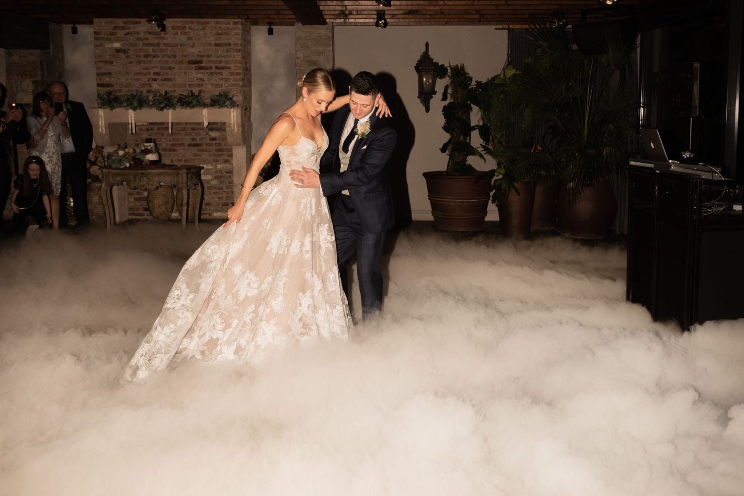 A groom affectionately holds his bride around the waist as he leads her to a dance floor. Low fog engulfs the ground around them.