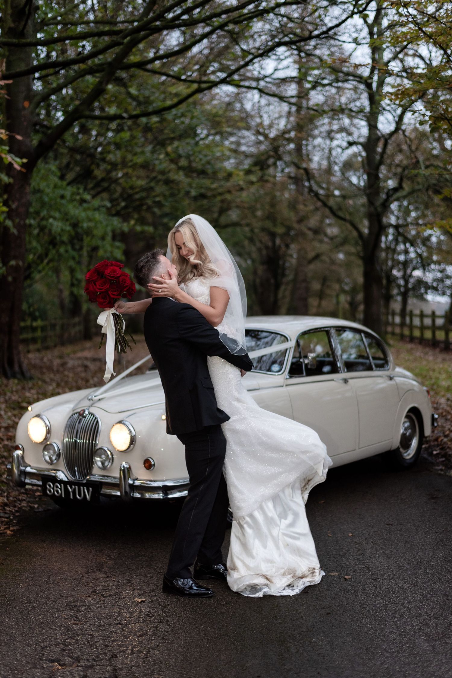 A bride smiles and holds her husbands face as he lifts her up into the air in front of a classic car on a tree lined road.