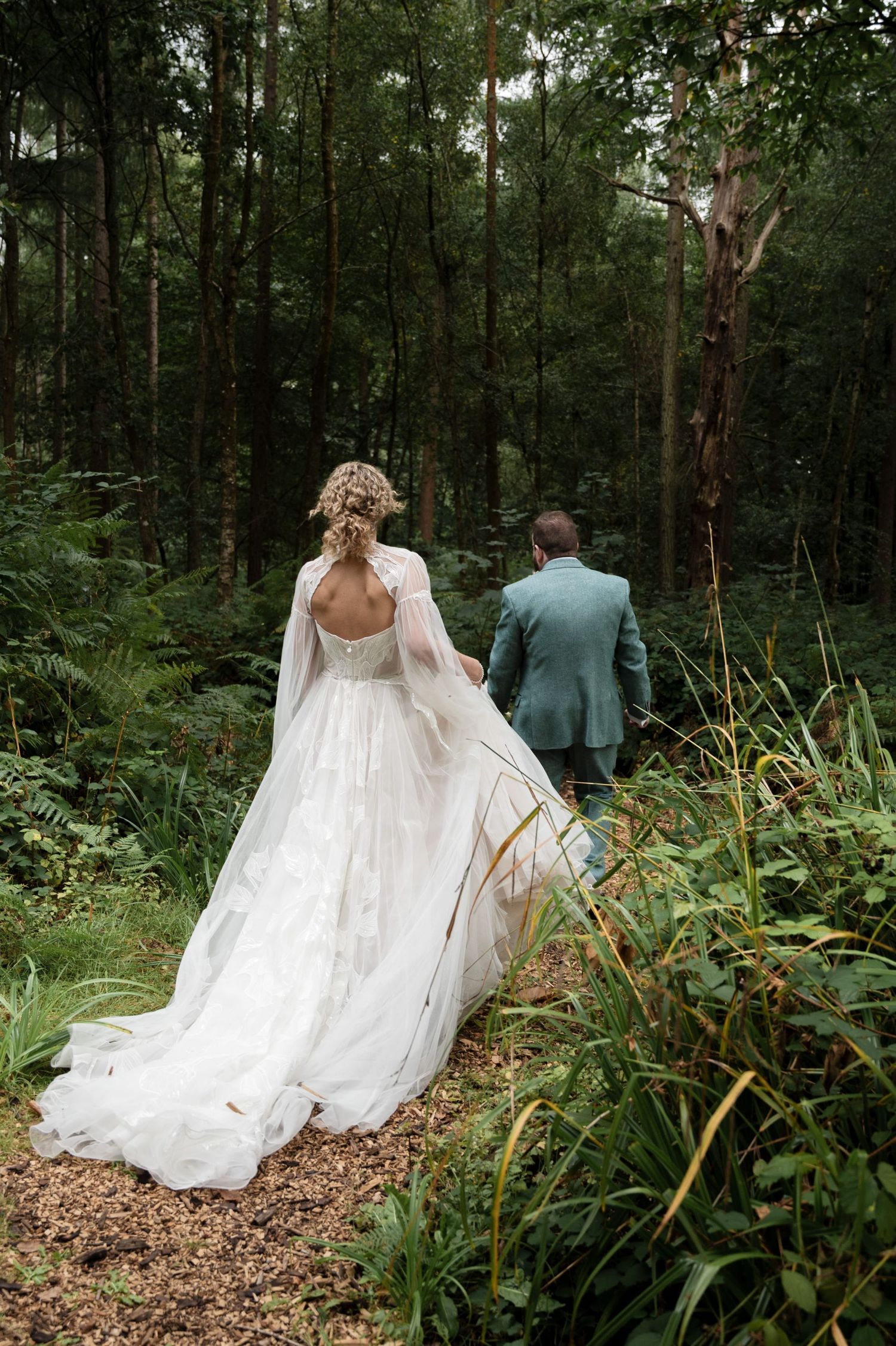Newlyweds walk into the forest, the brides skirts are long with a dramatic train which picks up the leaves as she follows her husband.
