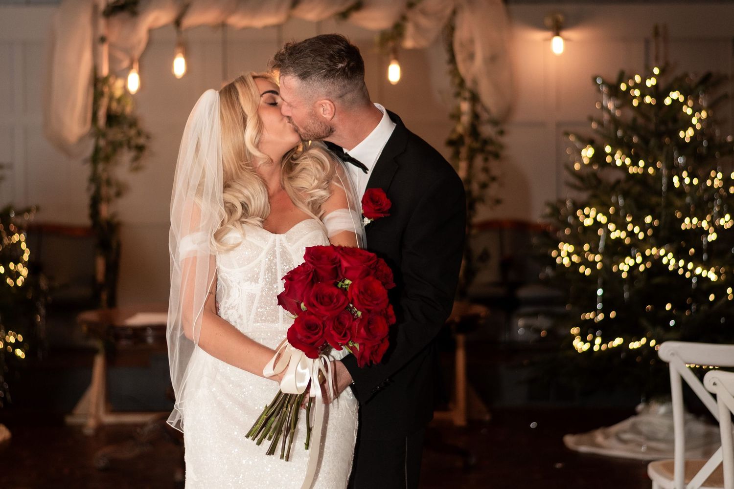 Bride stands in front of her husband and leans back to kiss him. She holds red roses and there is a glistening Christmas tree behind them.