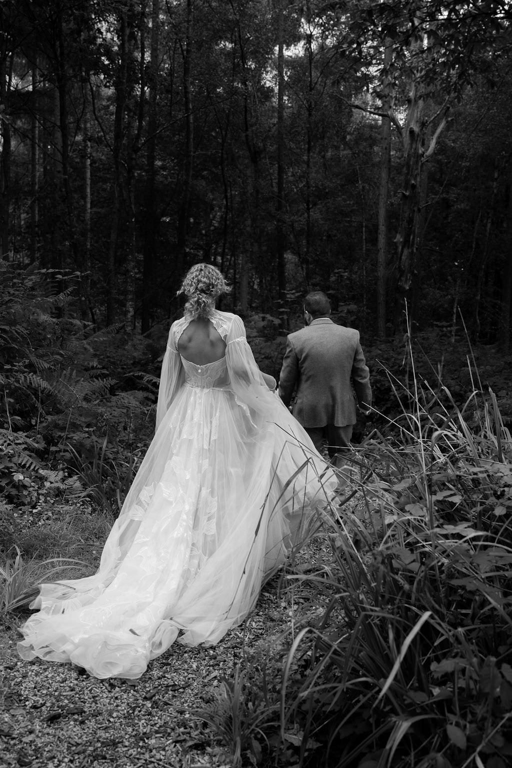 Bride in a gown with a long embroidered train walking through the undergrowth in a forest, the groom leads the way walking slightly ahead.
