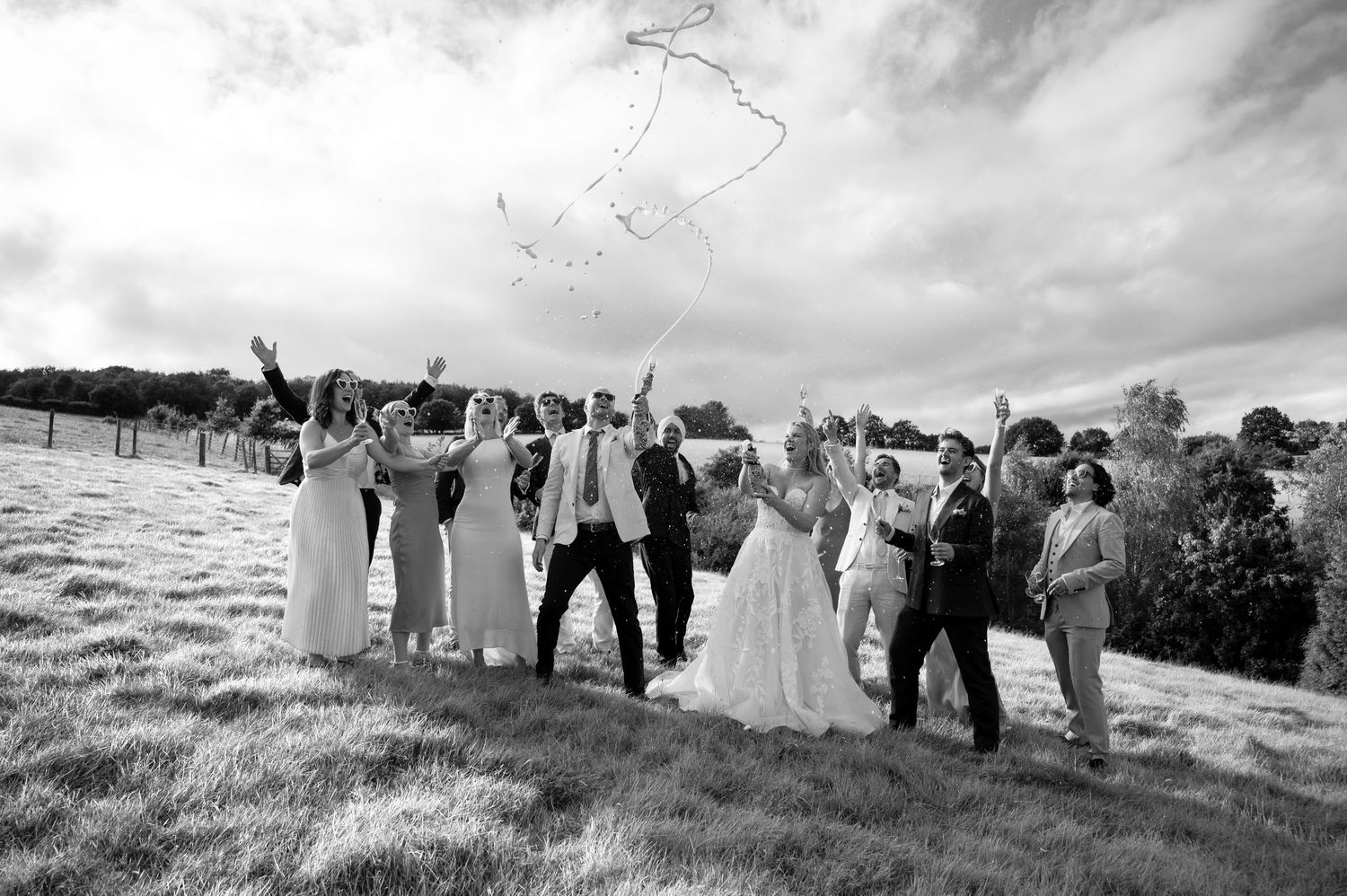 A bride and groom standing on a sloped hilltop laughing whilst spraying bottles of champagne high into the air with friends.