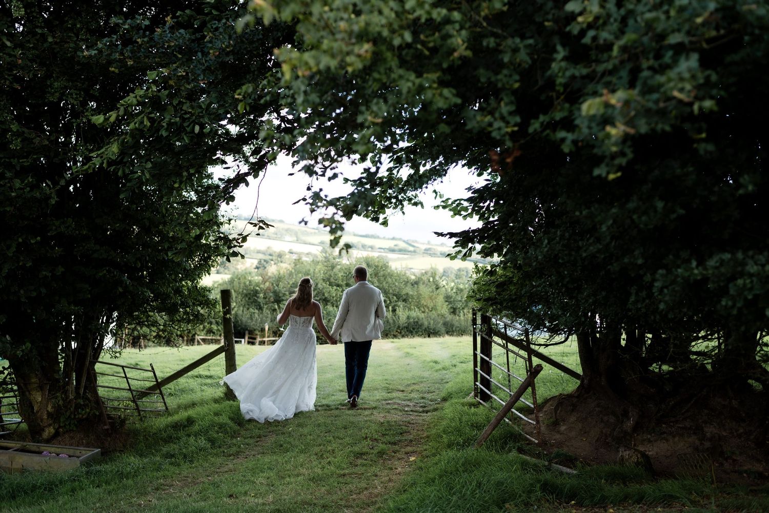 Groom leading his bride by the hand through tree lined farm gates down into a field below. The wind blows her gown behind her as she walks.