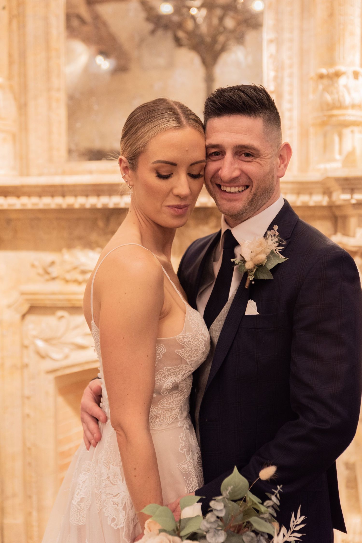 Groom pulling his bride in close so their heads are touching, he is smiling looking ahead whilst she is smiling looking down at her bouquet.