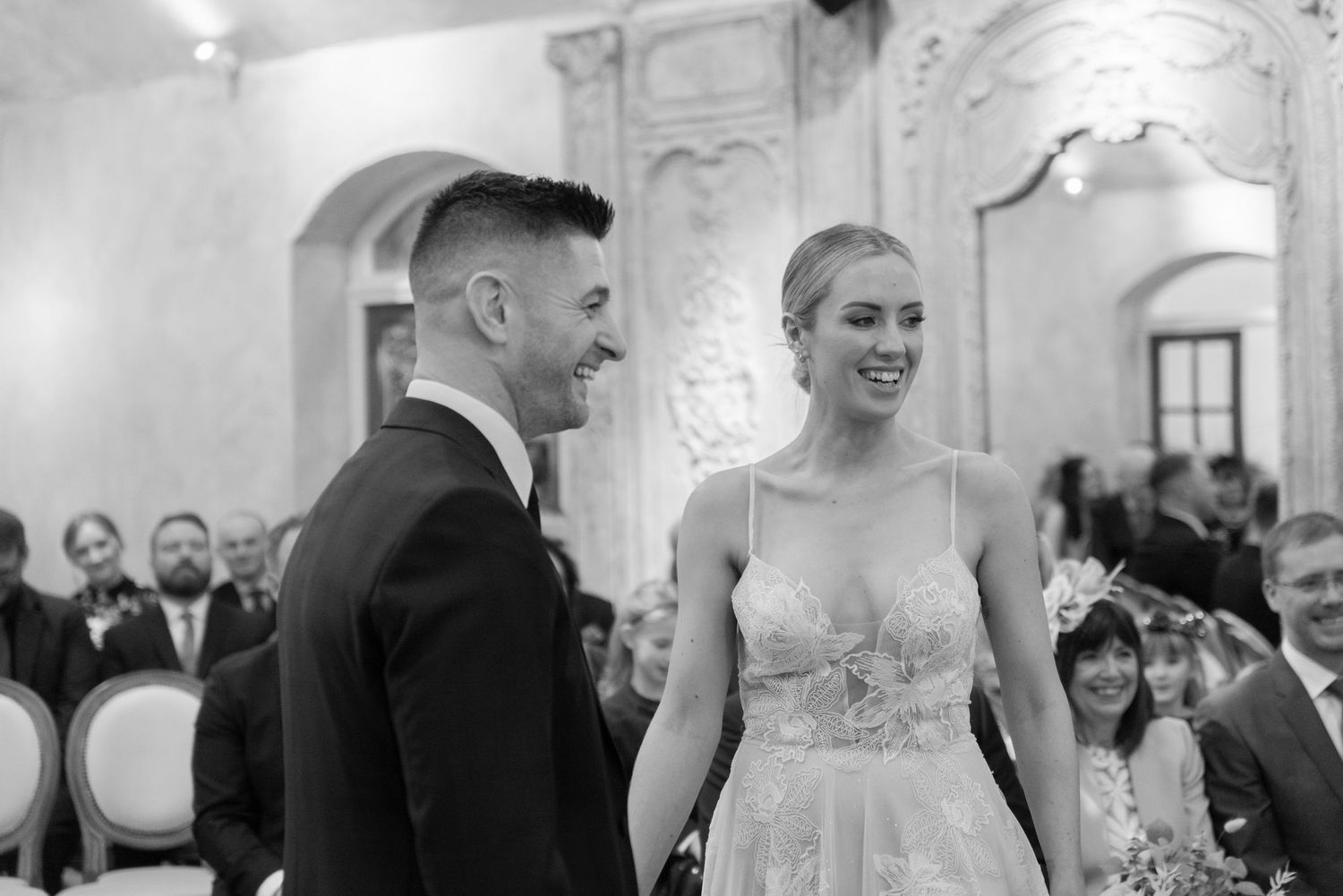 A bride and groom stand at the alter in a grand mirrored room whilst surrounded by smiling friends and family.