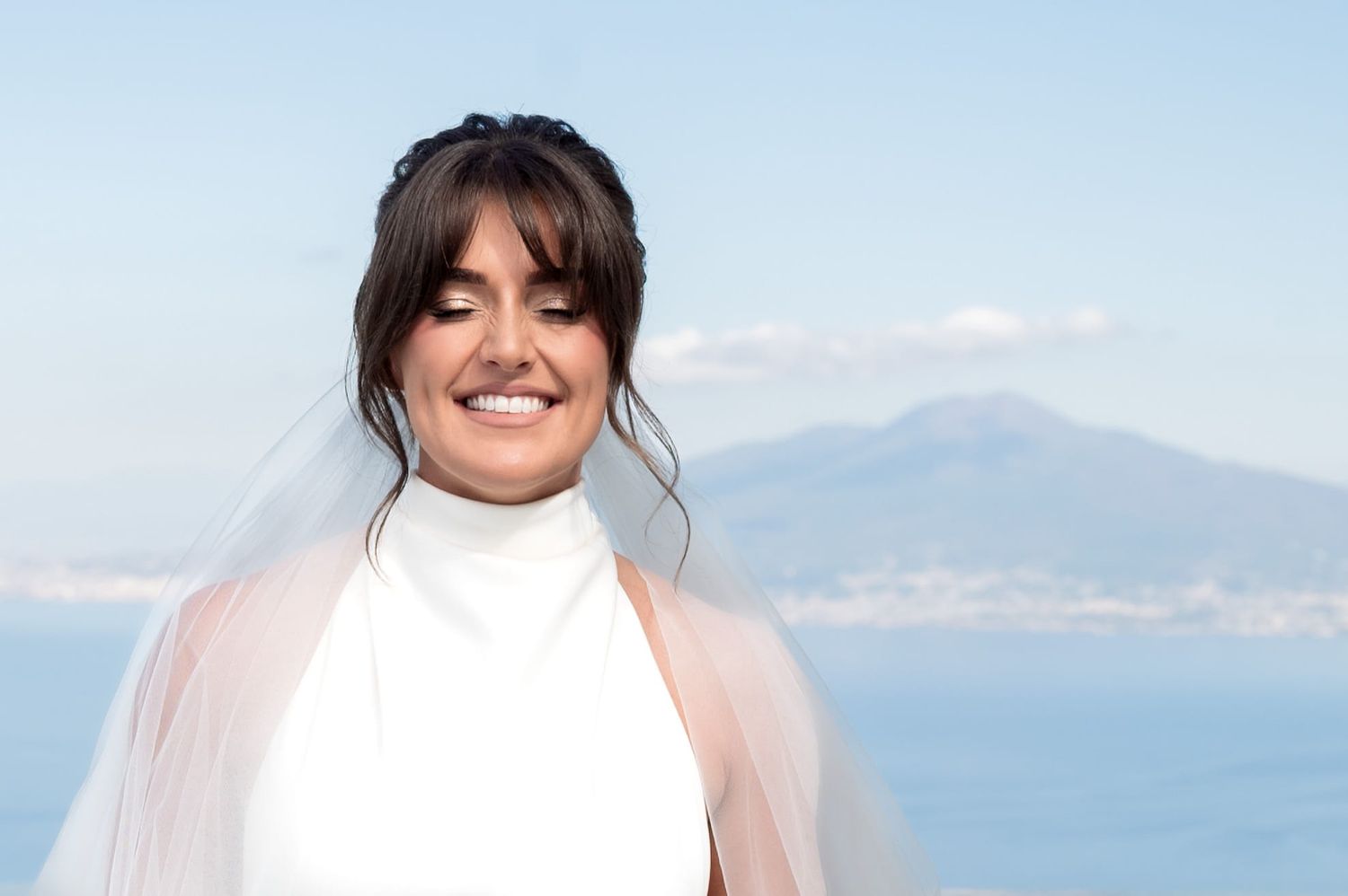 Woman in white dress and veil smiles on the coast overlooking Mount Vesuvius. The weather is bright and soft clouds surround the hills.