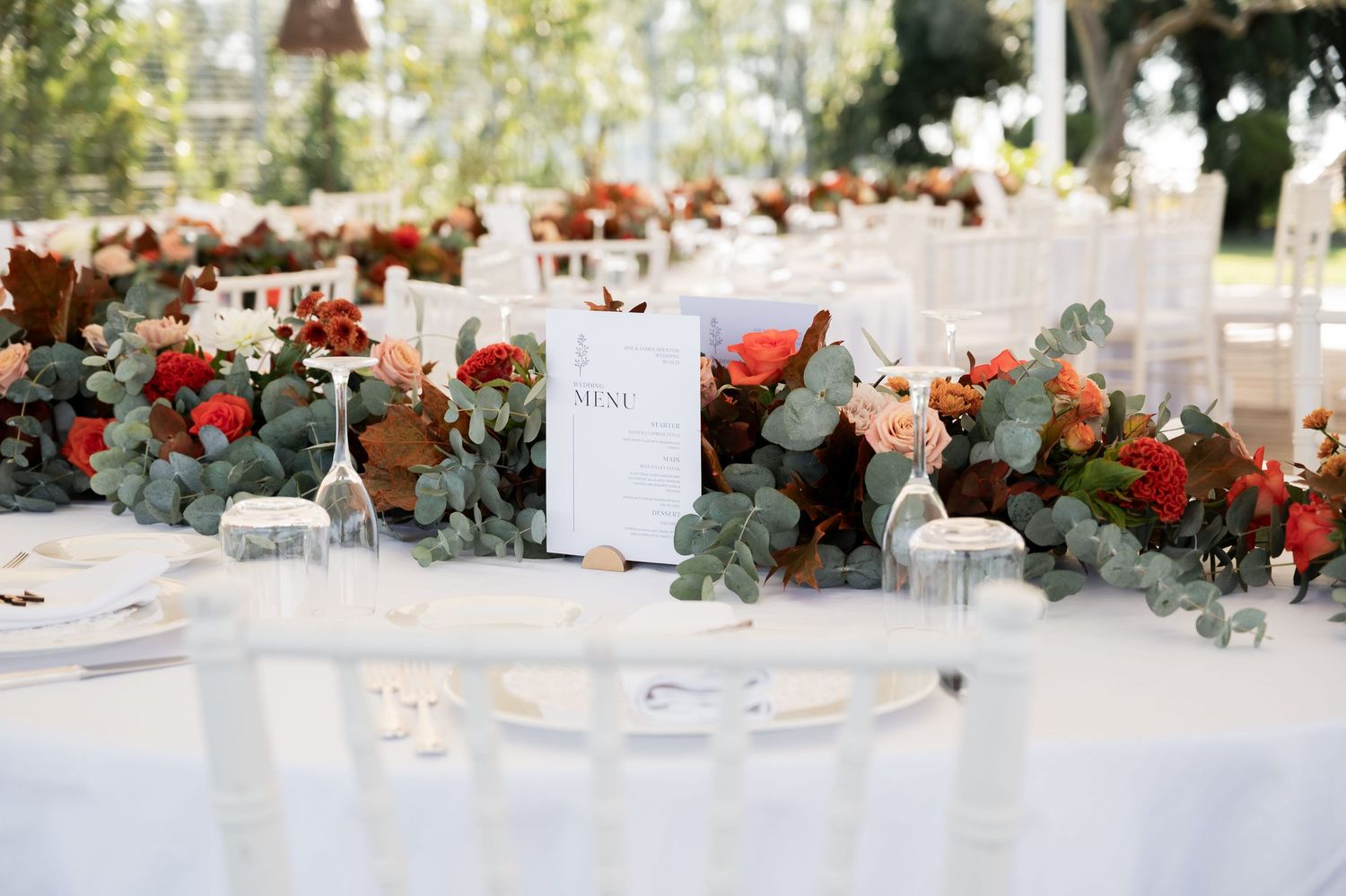 Blooming orange flowers dotted amongst green foliage, arranged on a large sweeping table which is set for a wedding breakfast.