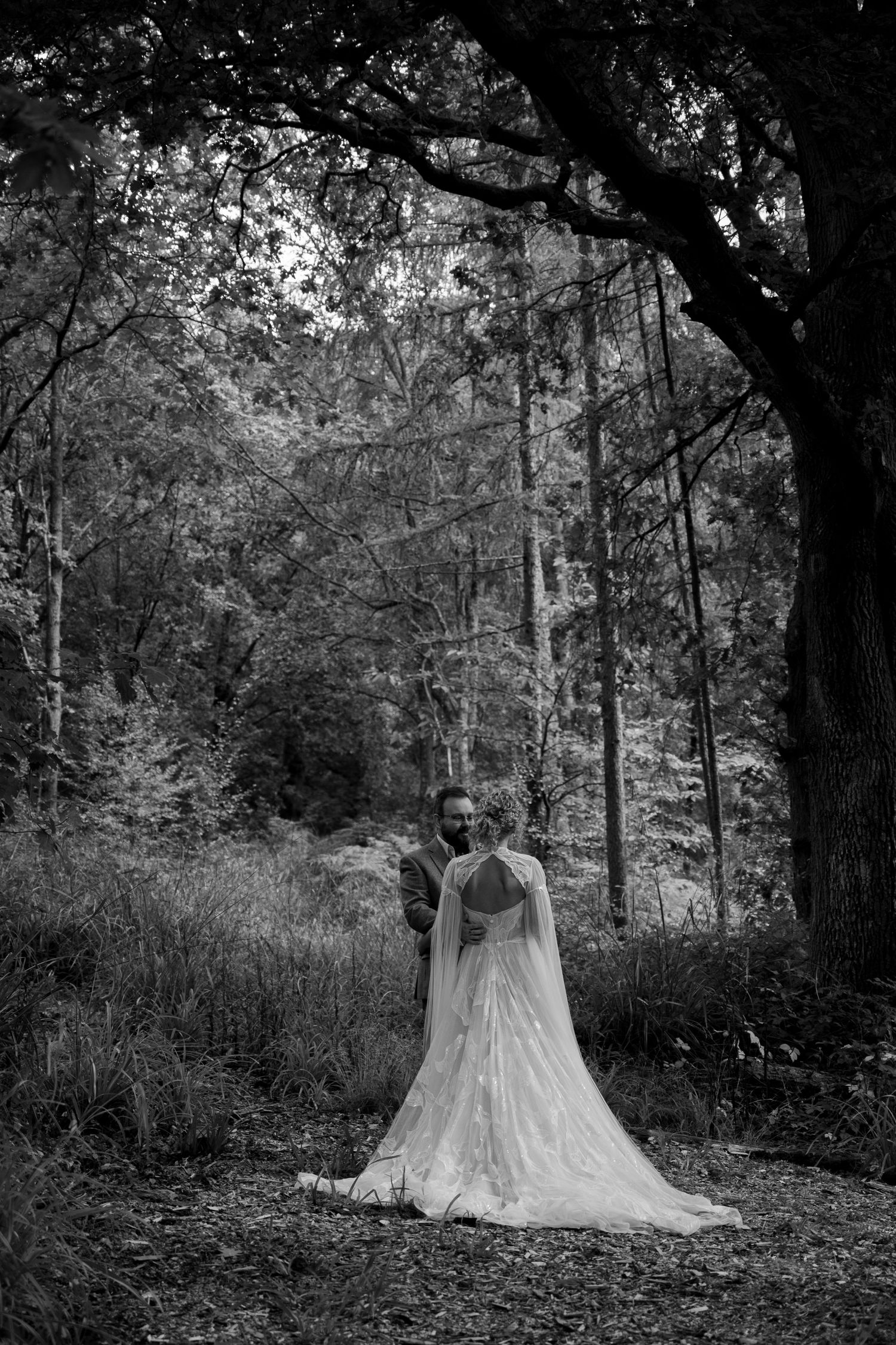 A bride and groom embrace in the forest surrounded by trees. The groom faces her, he has his arm around her waist and smiles.