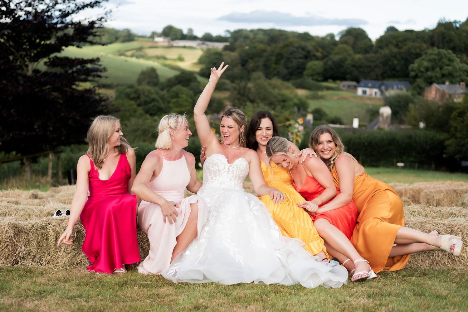 Group of women sitting on hay bales at the top of a hill laughing and hugging. They're wearing brightly coloured silk dresses.
