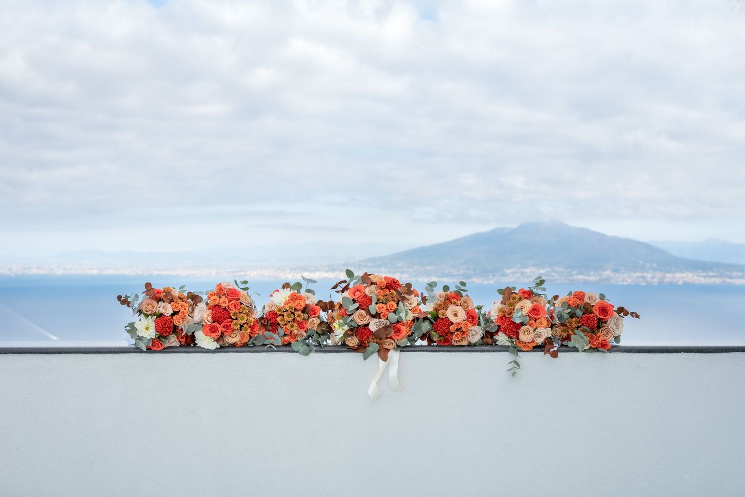 Row of orange and yellow floral bouquets neatly lined up on a wall, in the background are mountains and the sky is rippled with clouds.