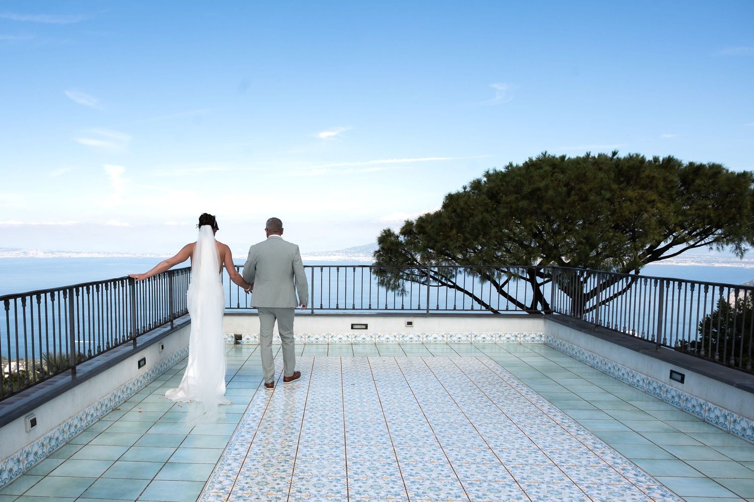 A bride and groom walking hand in hand along an intricately tiled rooftop terrace overlooking the Amalfi coast.