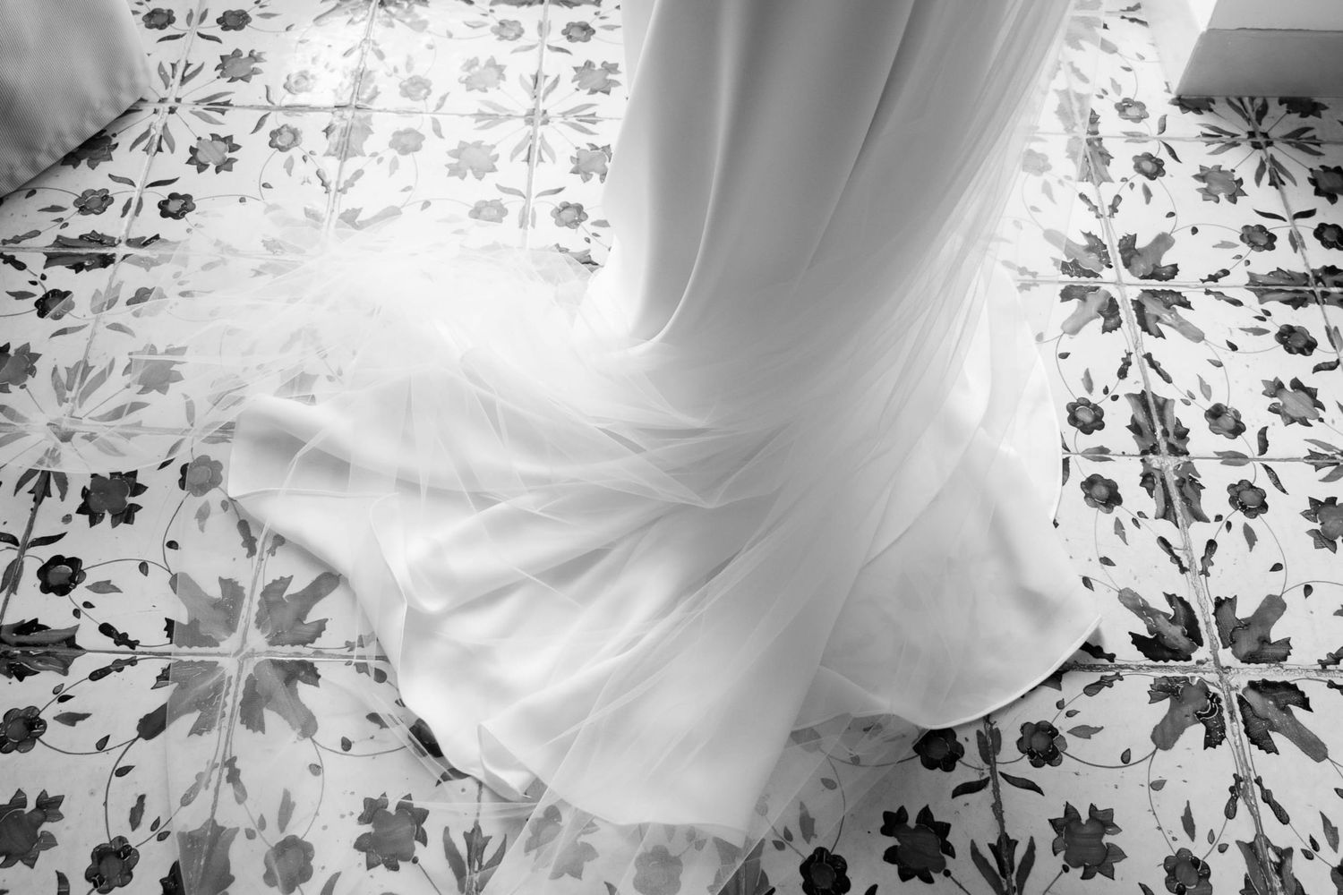 White wedding dress and train arranged in a swirl shape on an elaborately patterned tiled floor. Light from a nearby door seeps in.