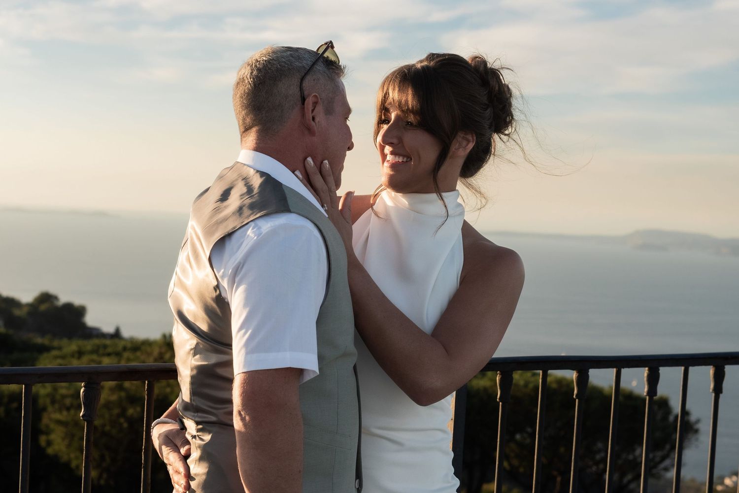 Couple standing on a high balcony with their arms around each other. They are by the ocean and their faces are highlighted by hazy sunshine.