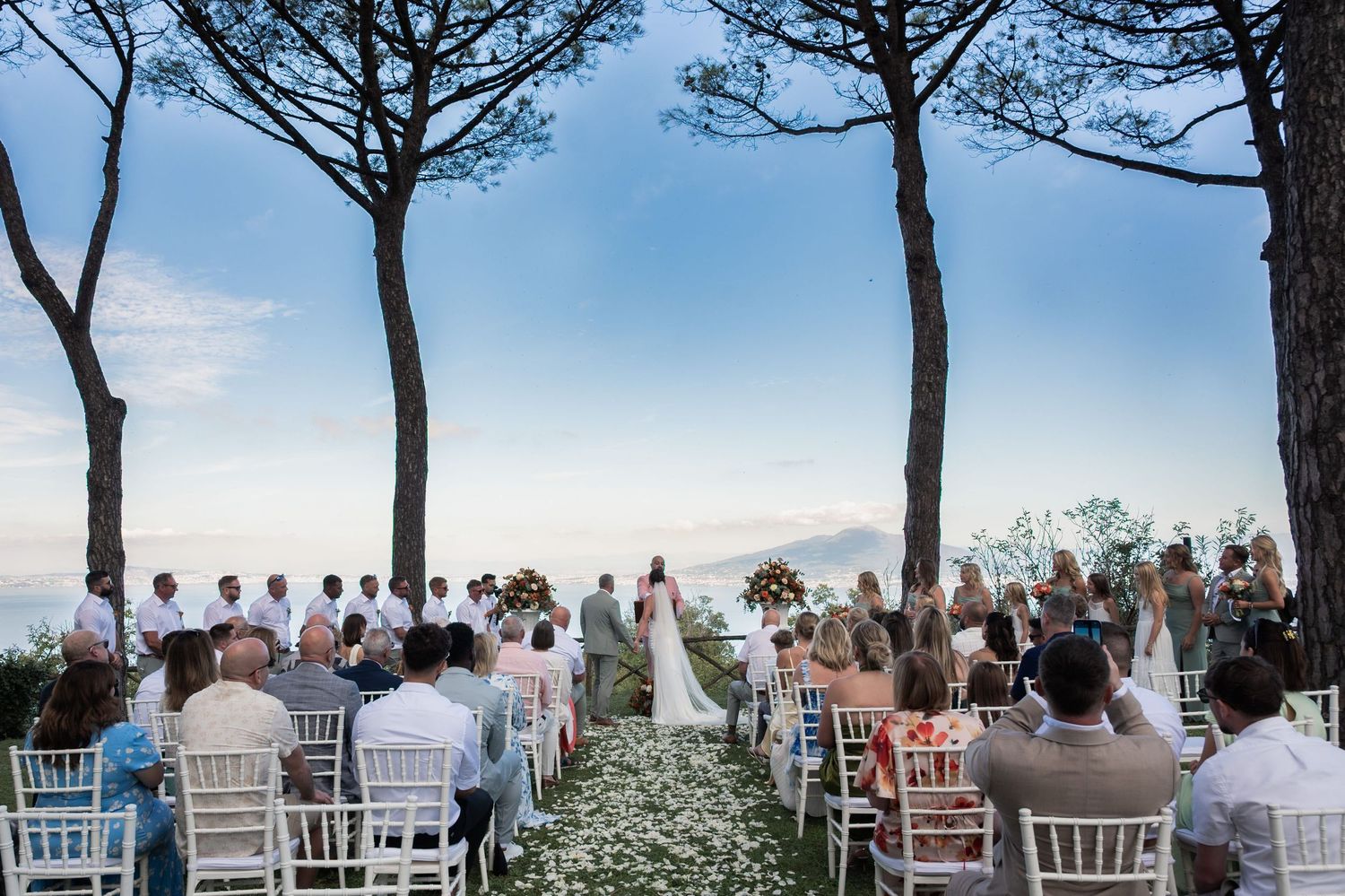 A wide angle shot of a wedding ceremony being held amongst tall trees on the Italian coast of Sorrento, overlooking Mount Vesuvius.