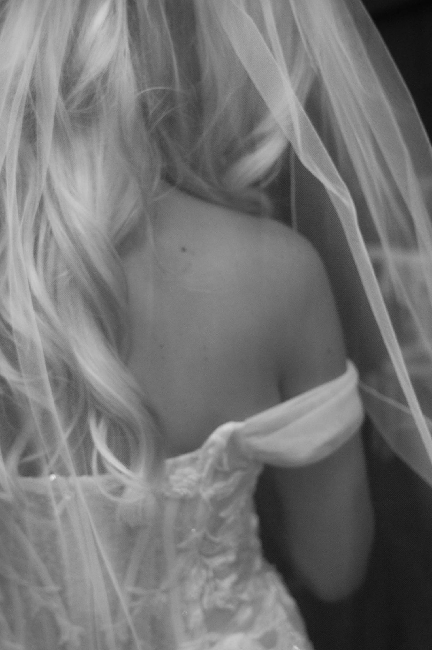 A bride photographed from behind, her blonde curls, long veil and lace sleeve falling gently over her shoulder.