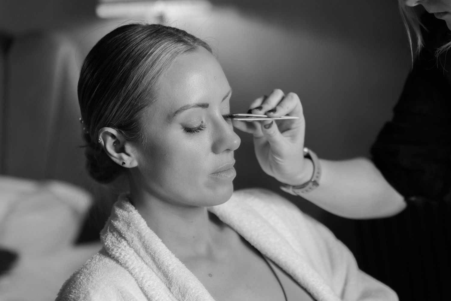 A woman wearing a bathrobe has false eye lashes applied by a makeup artist. The room is softly lit with a lamp.