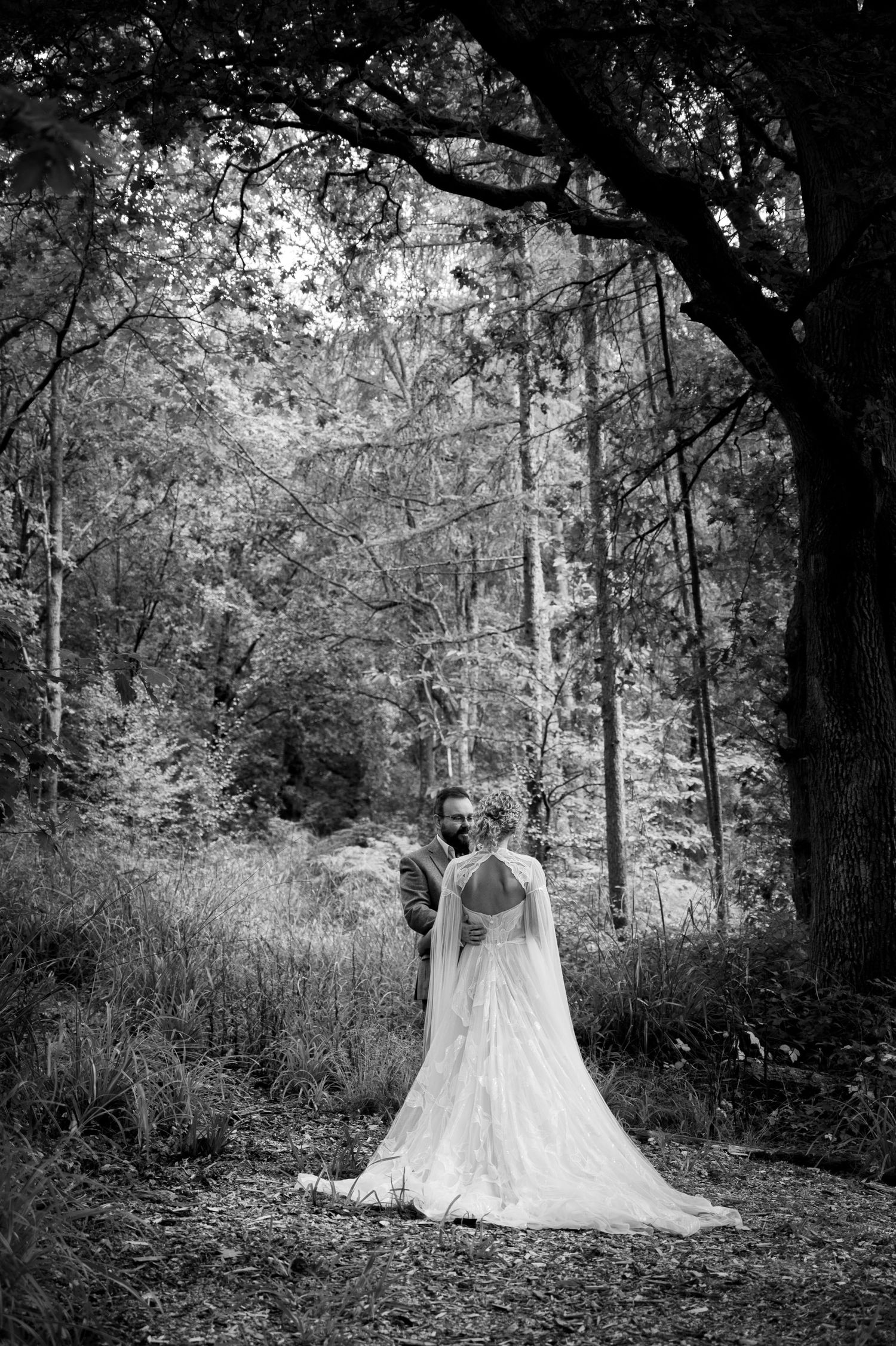 Happy couple stand under the cover of a large oak tree, the woman's face is obscured but the man holds her waist and smiles at her.