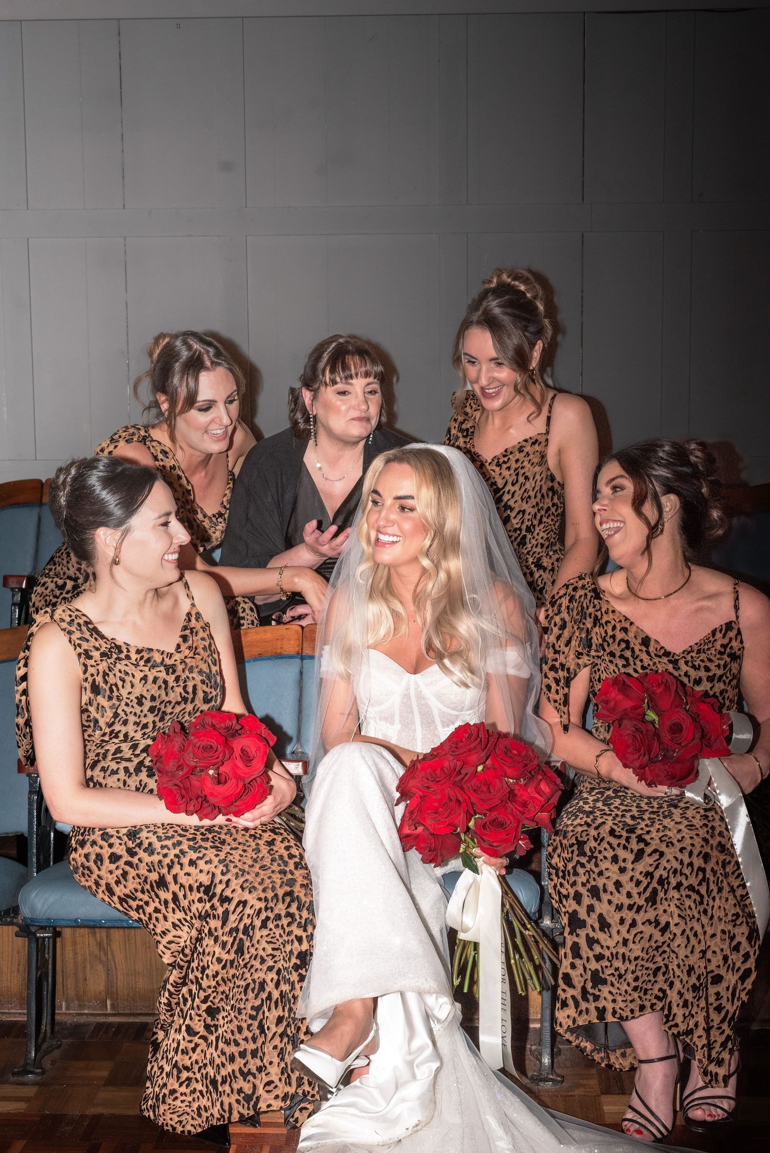A bride surrounded by her bridesmaids smiling, their dresses have a leopard print design and they are holding bouquets of red roses.