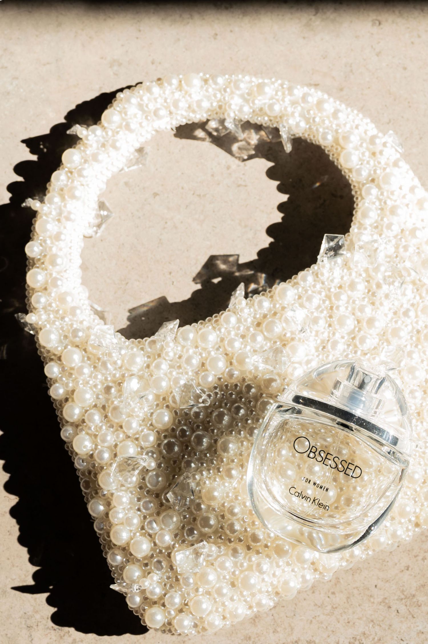 Bottle of Calvin Klein Obsessed perfume lay on a handbag decorated with jewels and pearls. Harsh sunshine illuminates the glass bottle.