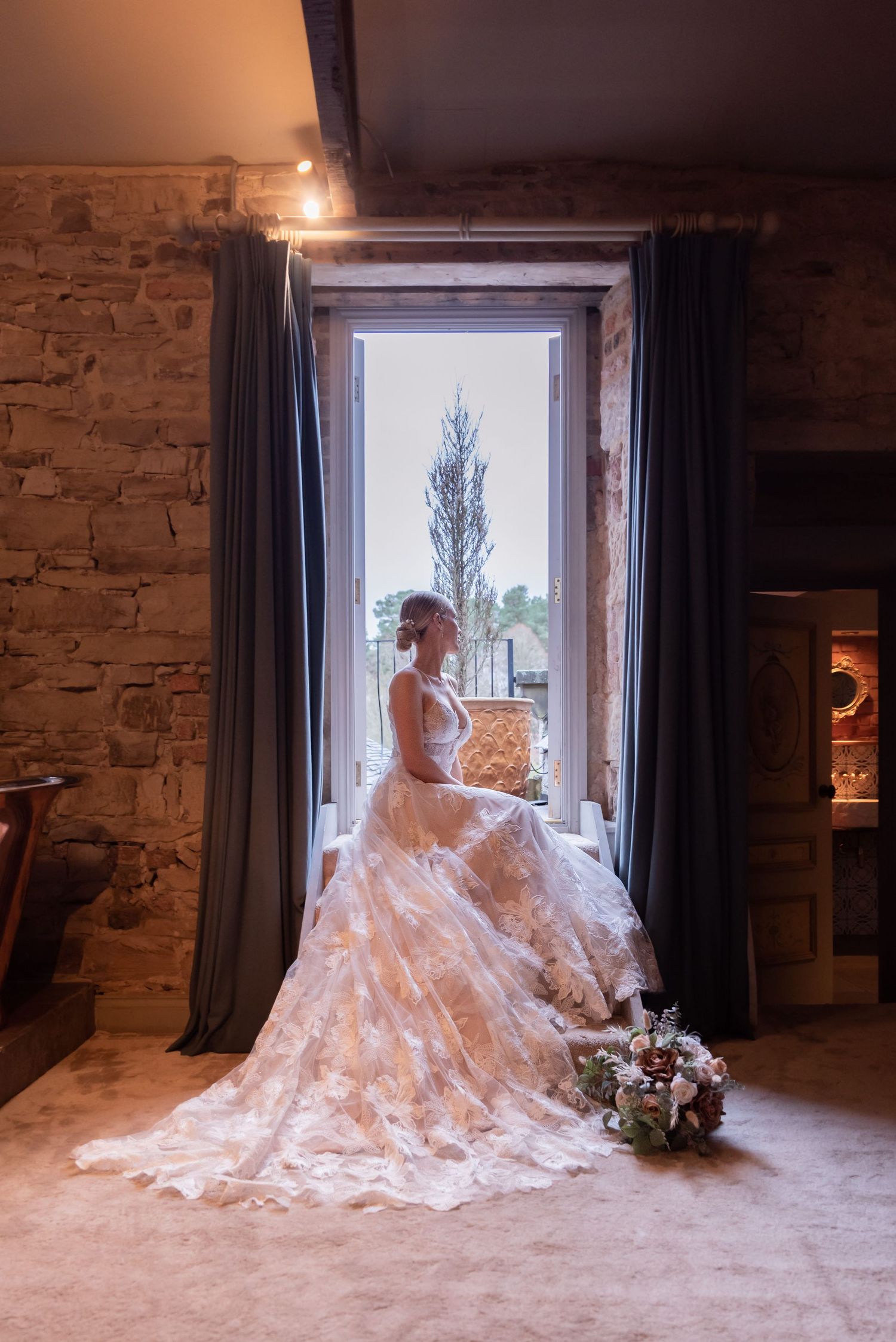 Woman in an intricate embroidered wedding gown with the train fanned out widely, sitting on a step peering out through an open door.