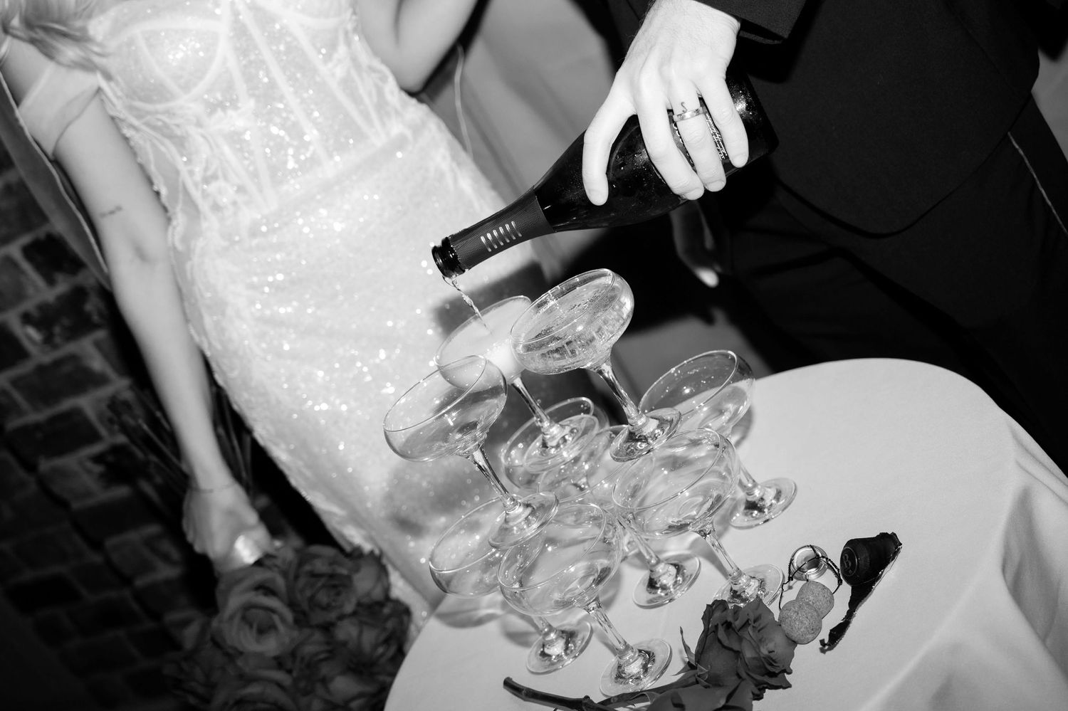 Champagne being poured onto a tower of glasses arranged in a pyramid. A rose and cork lie on the table and a bride stands behind.