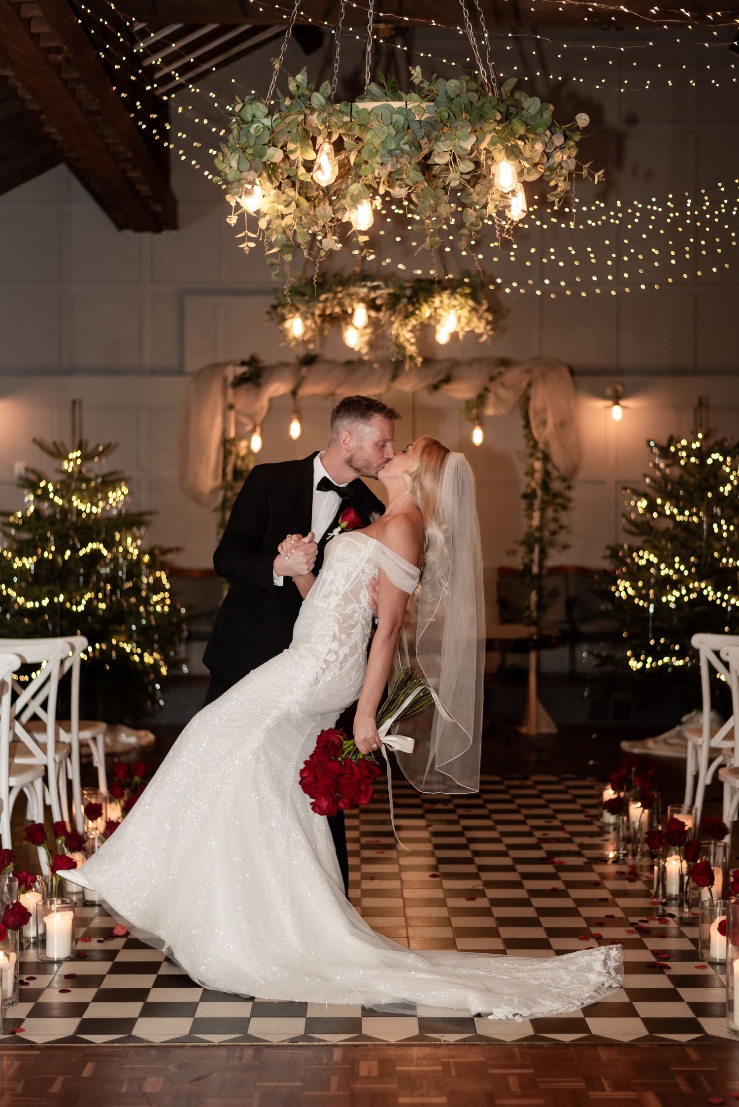 Groom dressed in black tie leaning his new wife back for a dip first kiss in a room lit by fairy lights and candles.