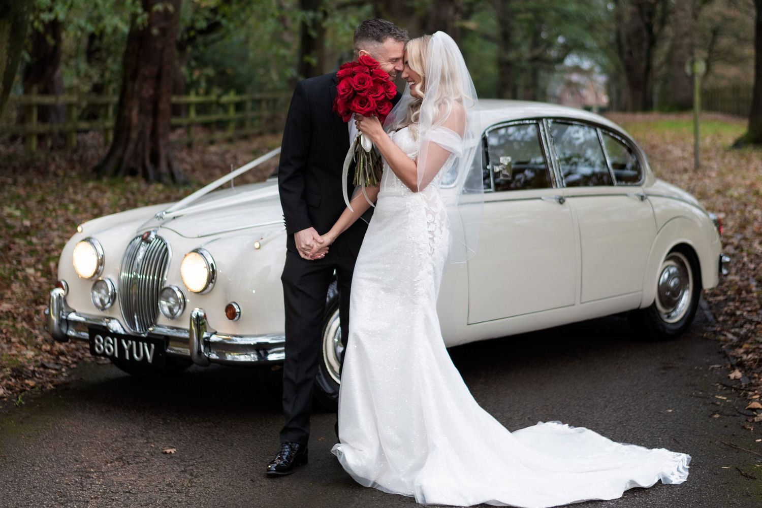 Bride and groom touching foreheads and clasping hands whilst they stand on a tree lined driveway in front of a cream coloured classic car.