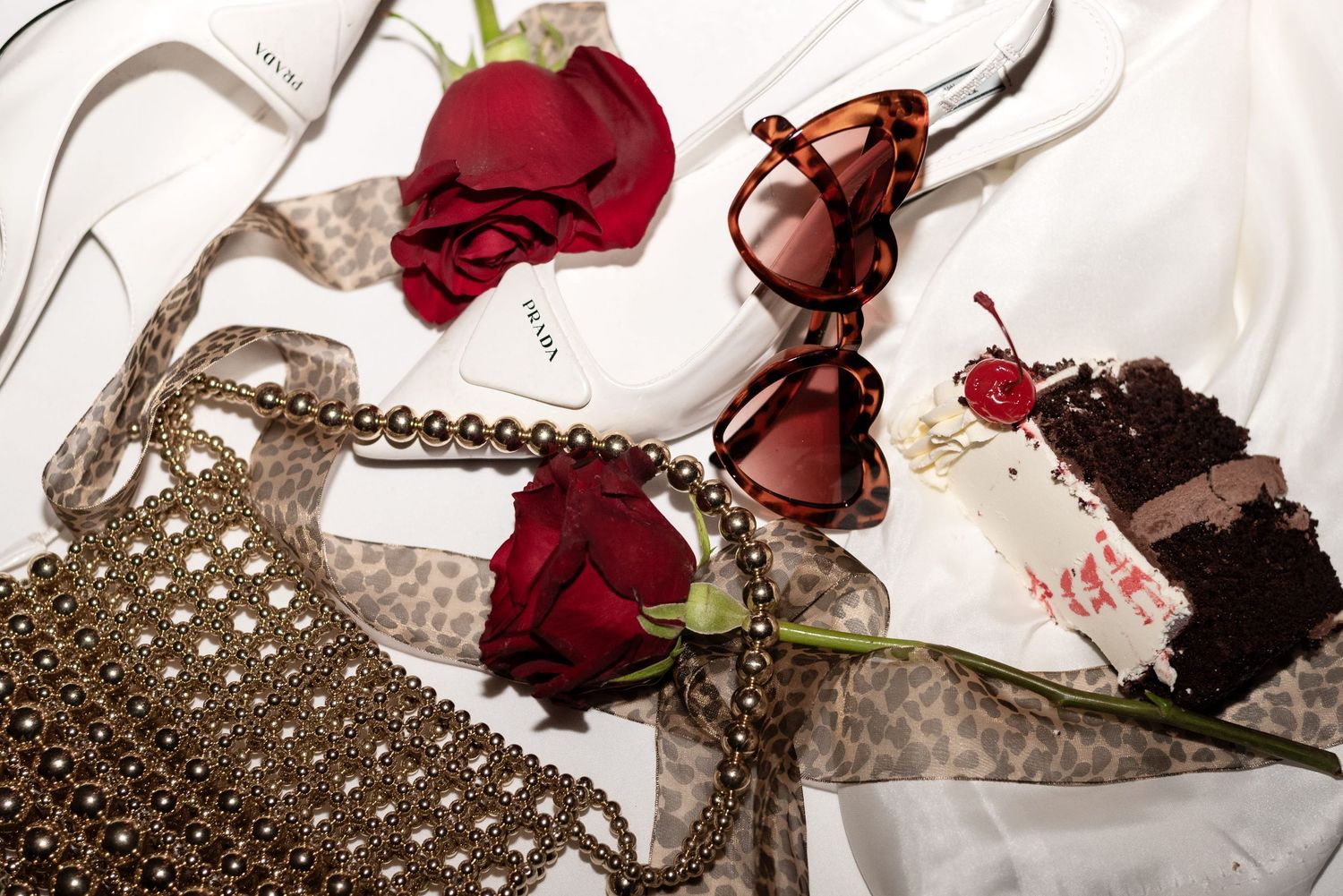 A pair of heart shaped sunglasses, a piece of cherry cake and a gold beaded handbag messily arranged on a table.