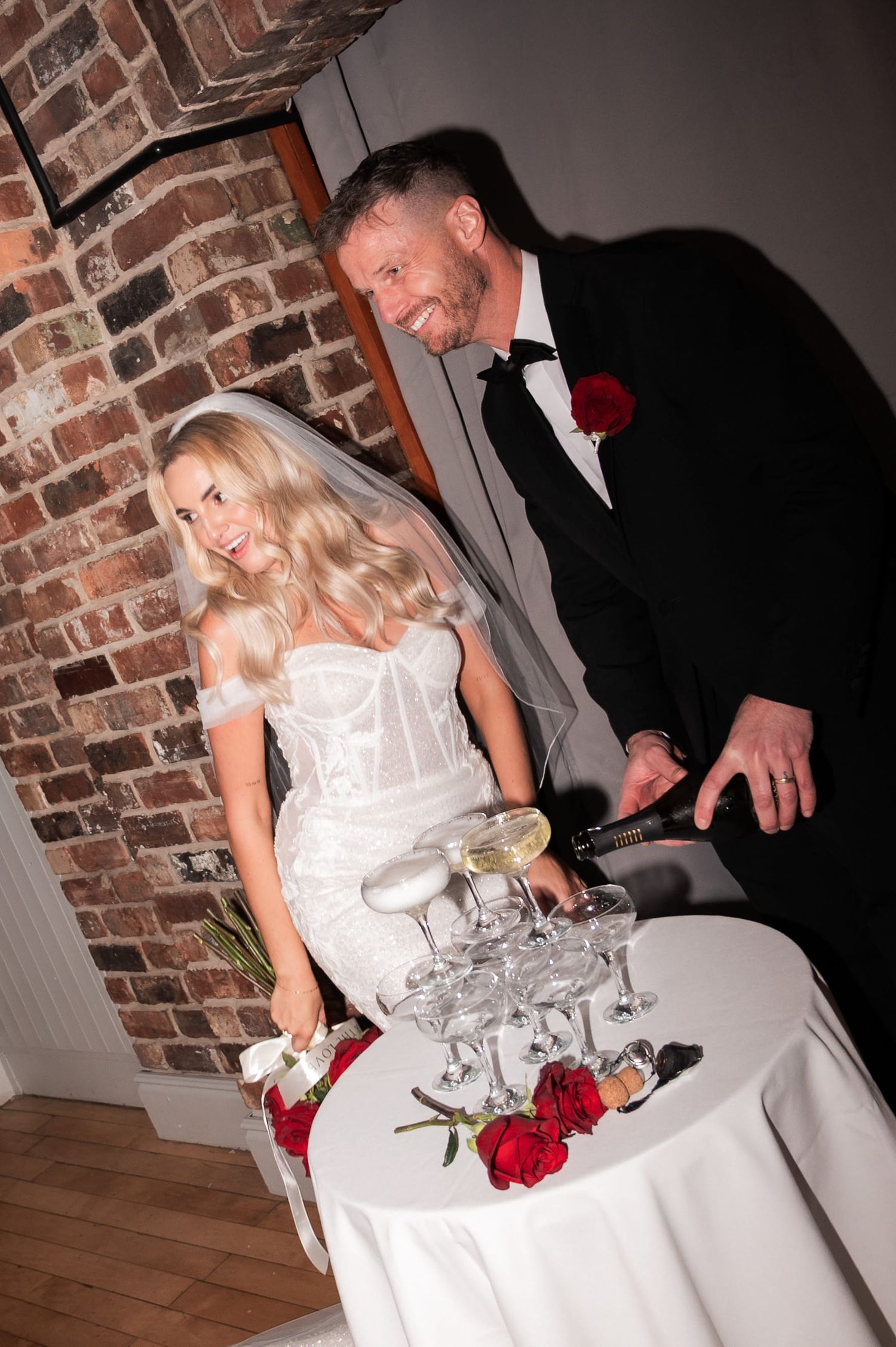 Bride and groom smiling whilst standing at a table decorated with a tower of glasses. The groom holds a bottle of champagne ready to pour.