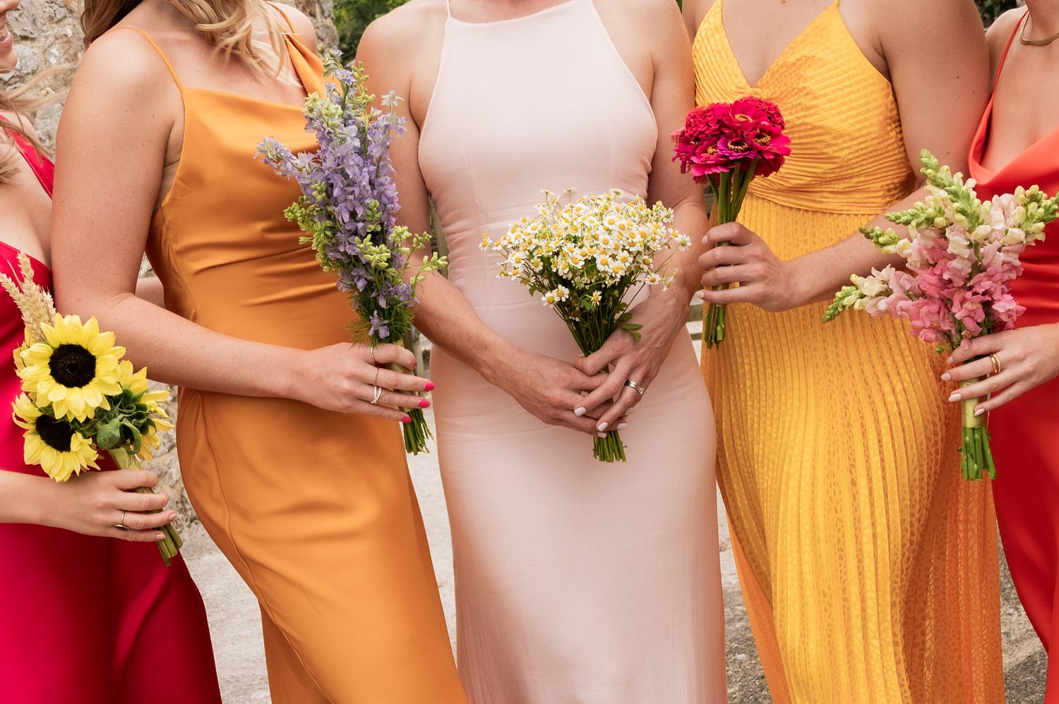 Five women in multicoloured dresses hold wildflower poseys at their waists. The posey's are purple, red, yellow, pink and purples.