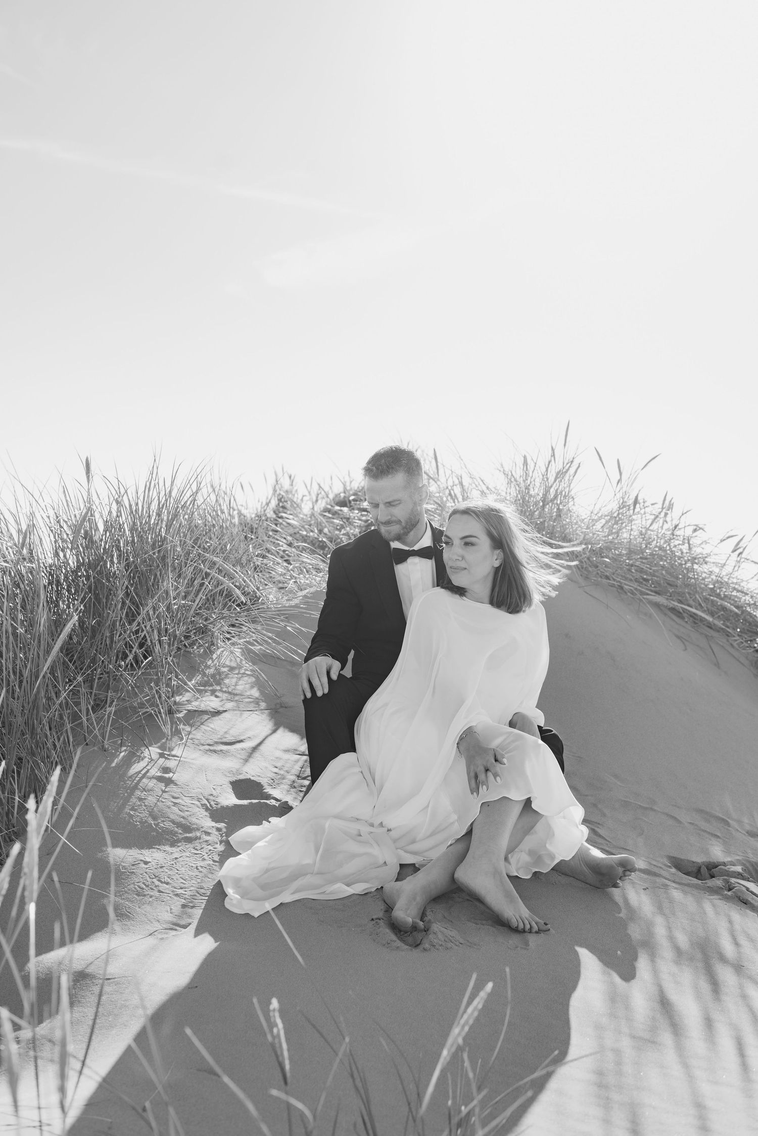 A man in a suit and woman in a flowing white dress sit on top of a sand dune surrounded by long grasses.