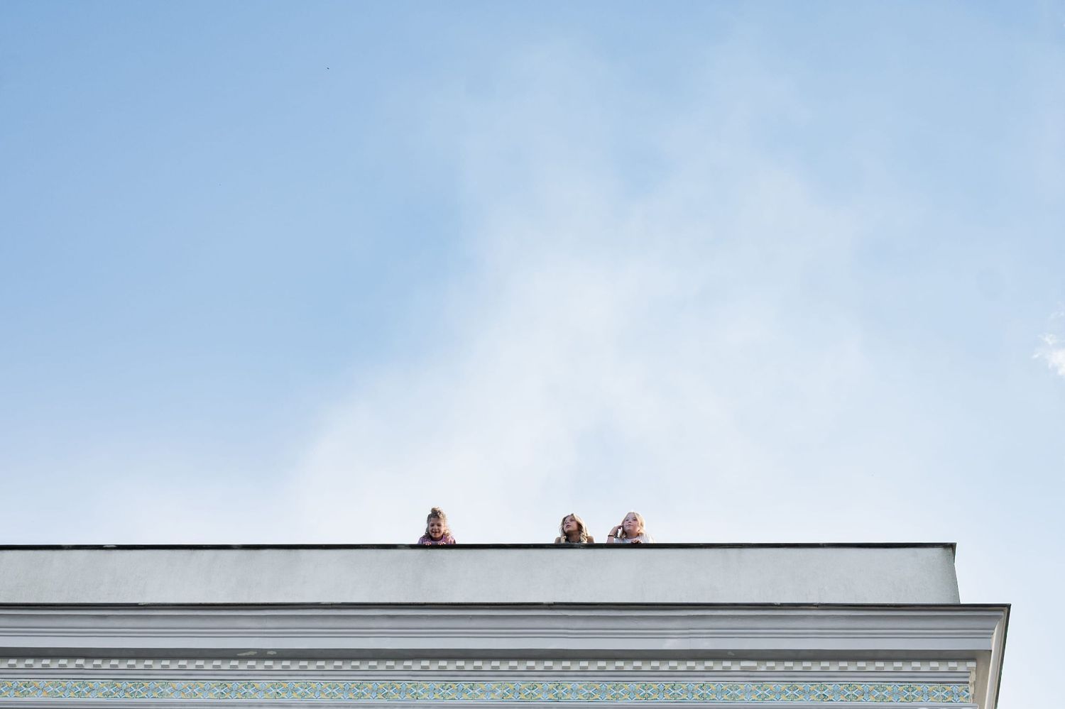 Three young children peer over the edge of a roof top balcony. White fluffy clouds drift above them emphasising how high up they are.