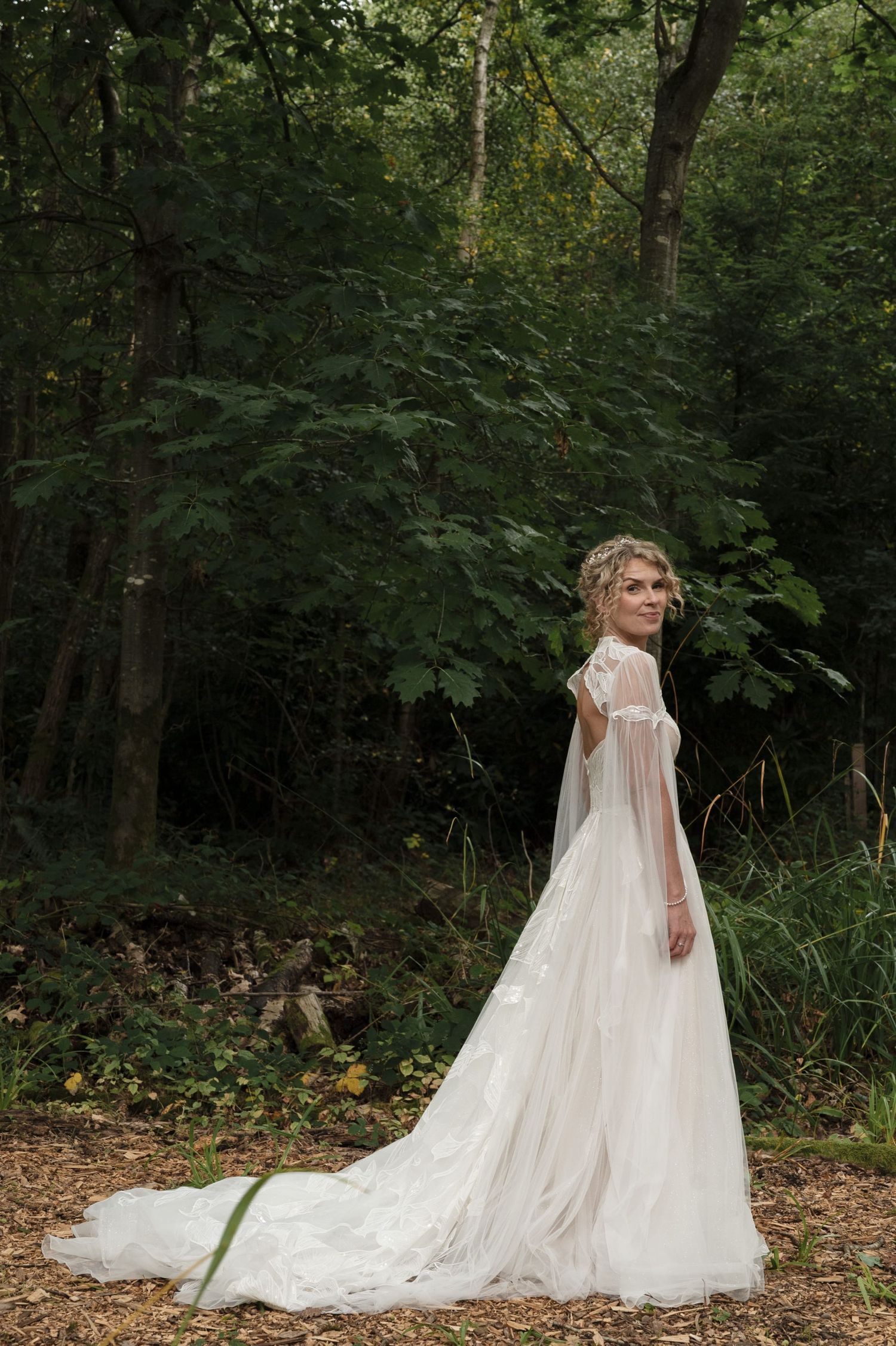 Blonde curly haired bride in side profile looking to camera, wearing a white patterned gown with long draped sheer sleeves.