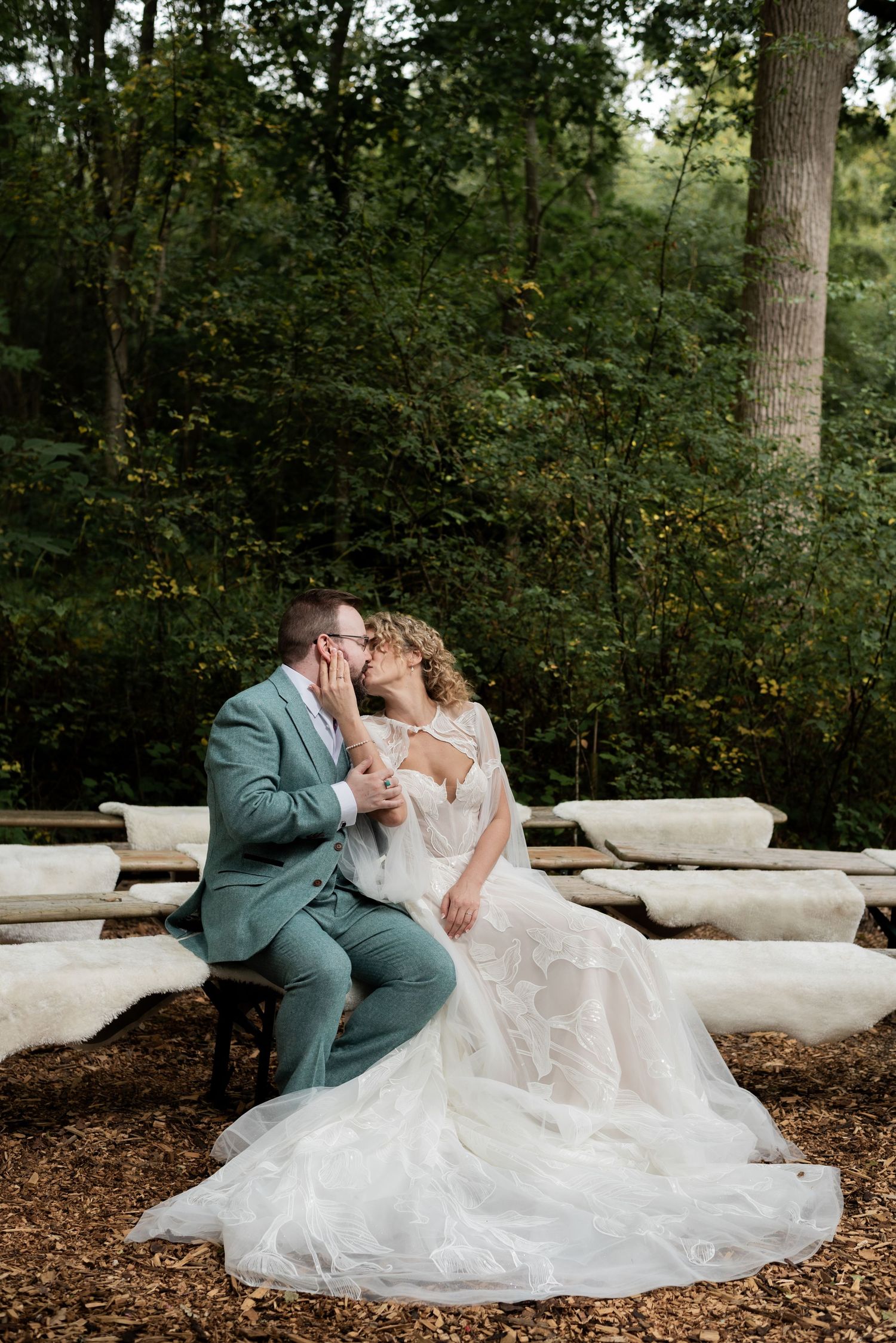 A groom wearing a sage green suit kisses his bride, she wears a white gown with long white train. They sit on a blanket covered bench.