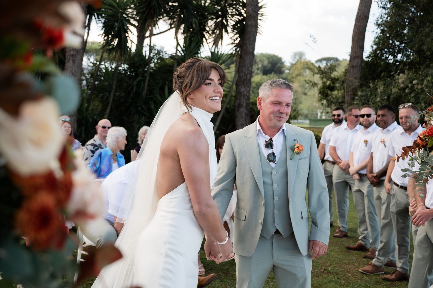Married couple stand at the altar of their outdoor wedding, beside a decorative floral structure. Several groomsmen smile beside them.