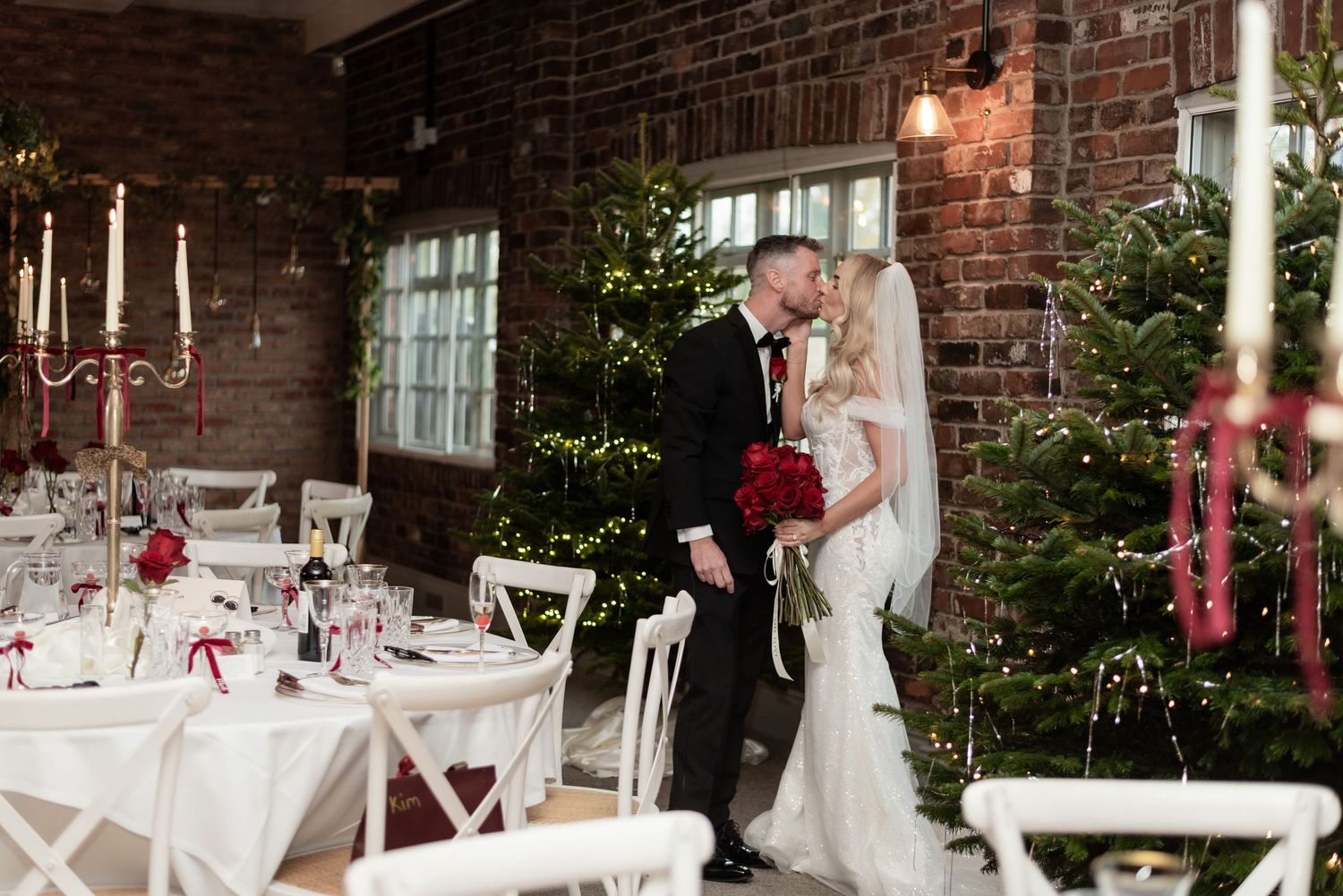 A married couple kissing in a brick walled dining room, the room is decorated with Christmas trees, tall candle sticks and red roses.