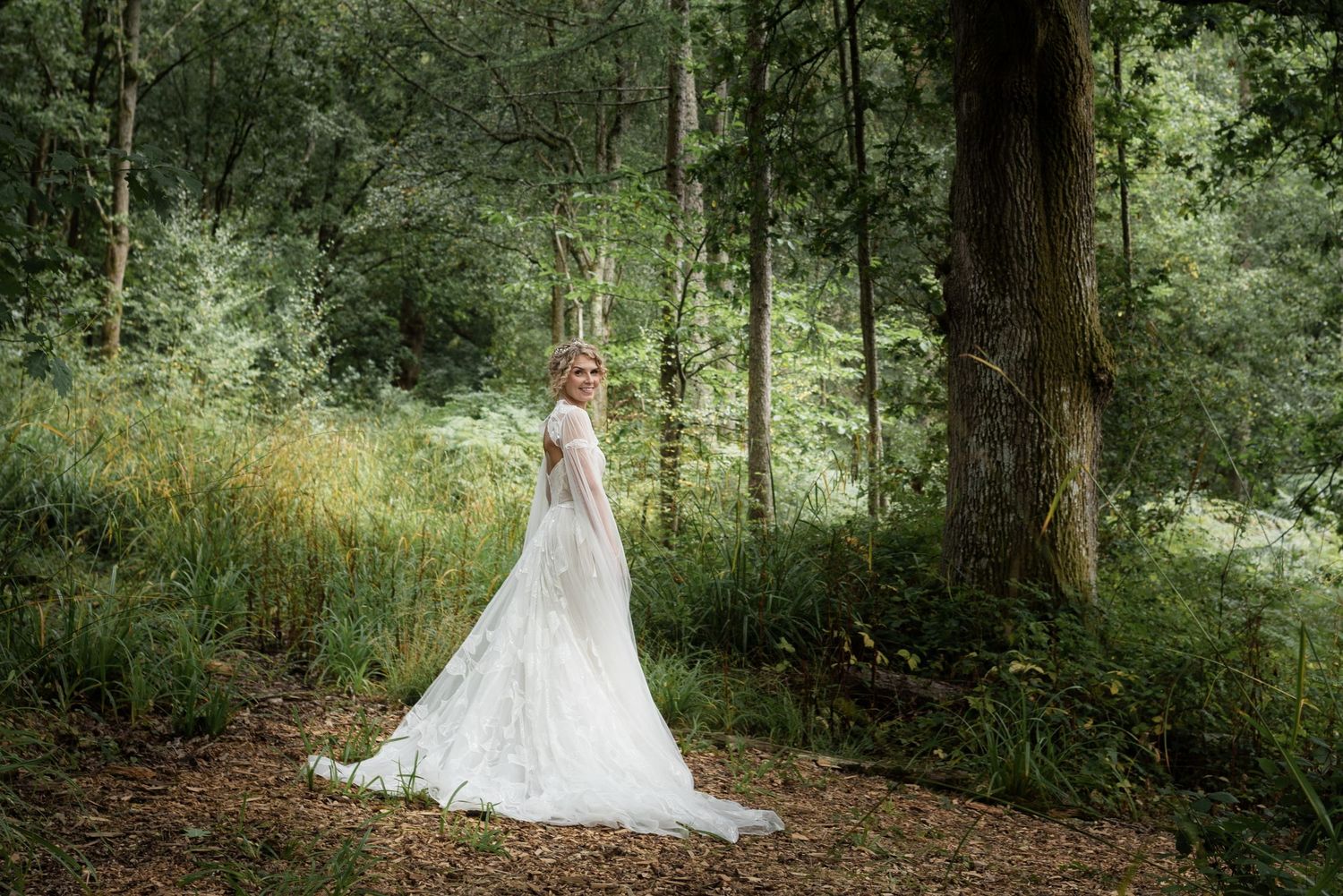A blonde curly haired woman stands deep in the forest wearing a large opulent white gown. Dense shrubbery and tall grasses surround her.