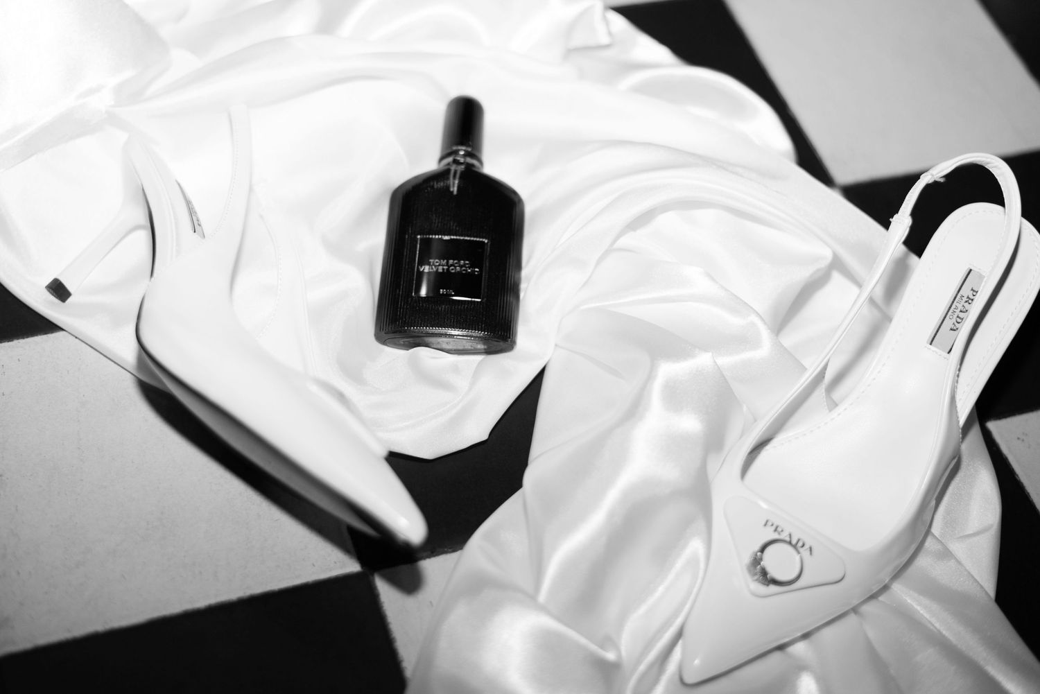 White silk sheet strewn across a black and white tiled floor. On top of the sheet lies a perfume bottle, white stiletto heels and a ring.