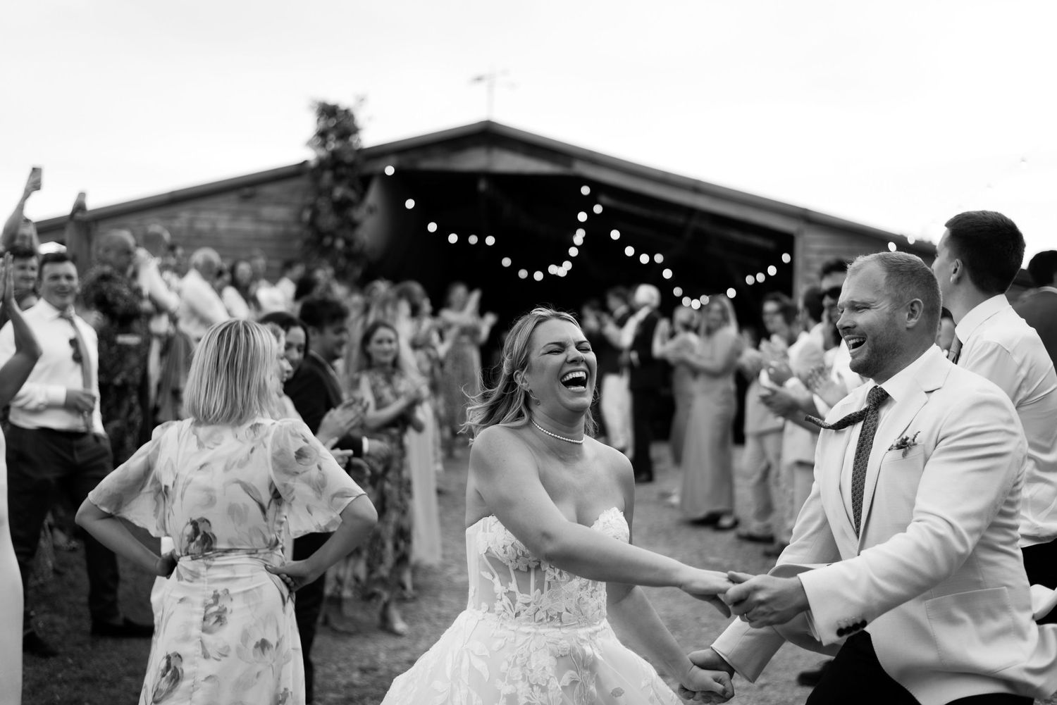 A bride and groom Céilié dancing hand in hand down a hill surrounded by friends. There is a fairy lit barn in the background.
